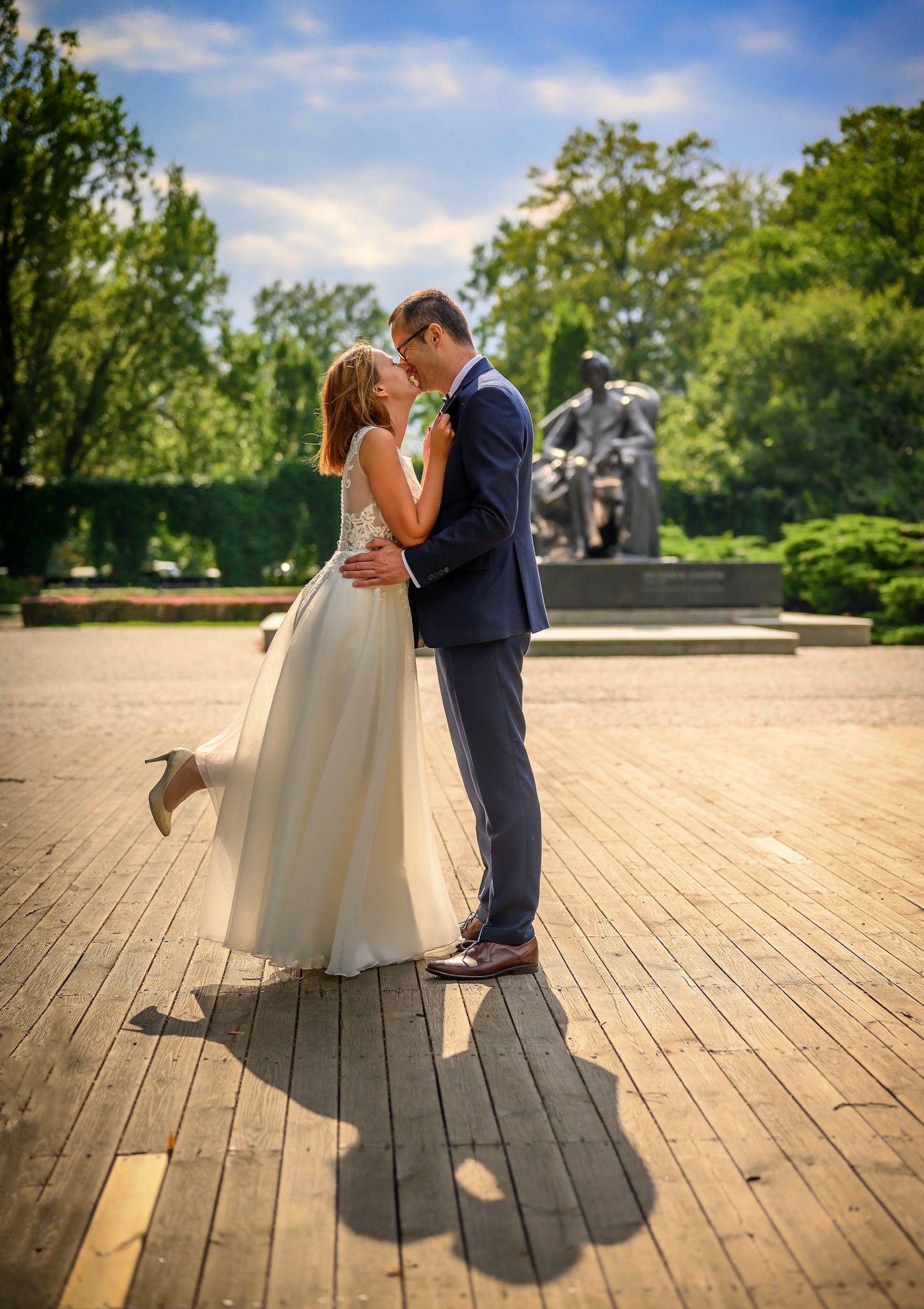 Para młoda całuje się na drewnianym podeście w parku, w tle pomnik i zieleń, panna młoda w białej sukni z koronkową górą i beżowych szpilkach, unosi jedną nogę.