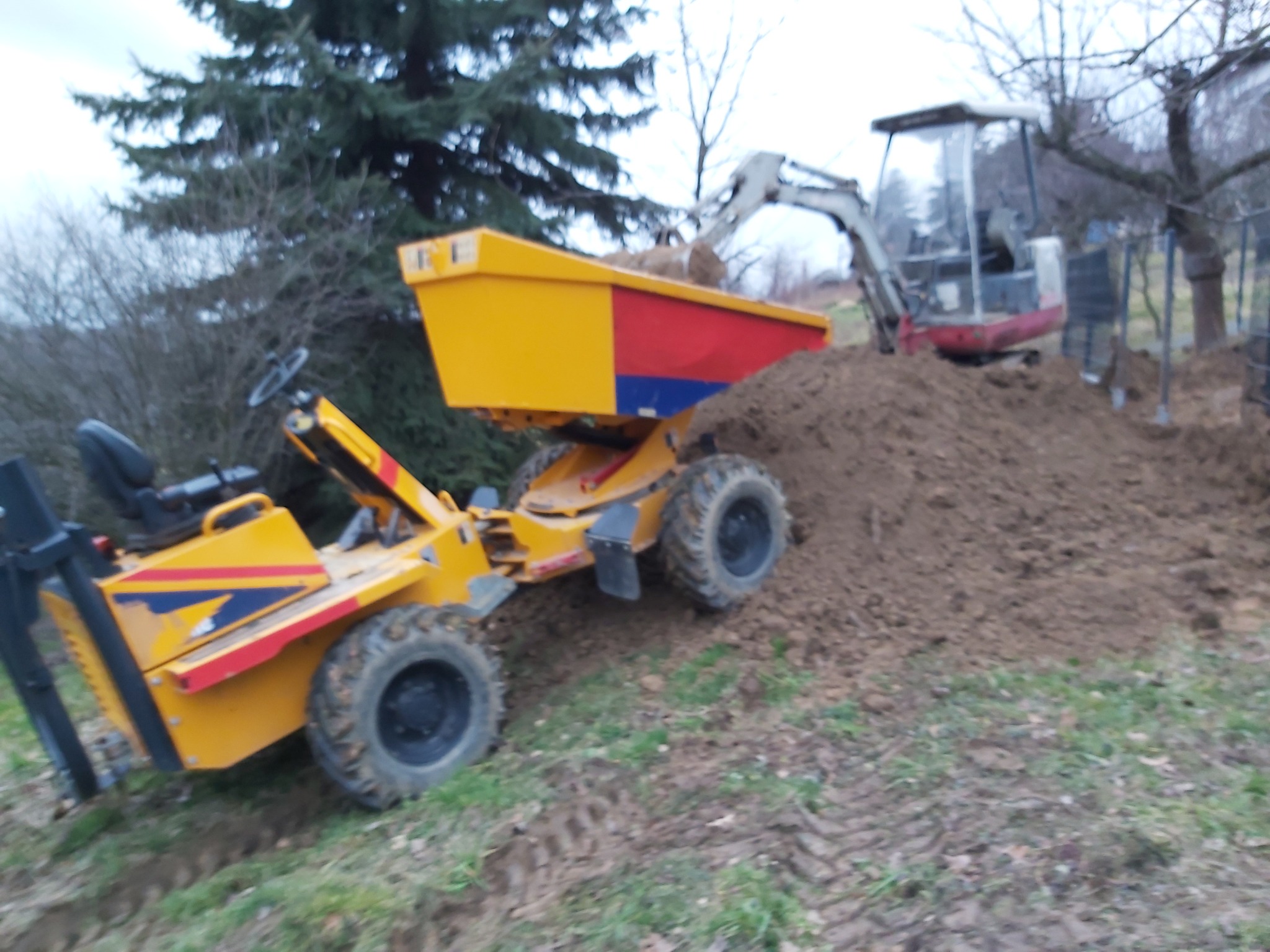 Żółto-czerwono-niebieska wywrotka na kołach z ładunkiem ziemi, obok koparka w trakcie pracy, teren budowy z drzewami w tle.
