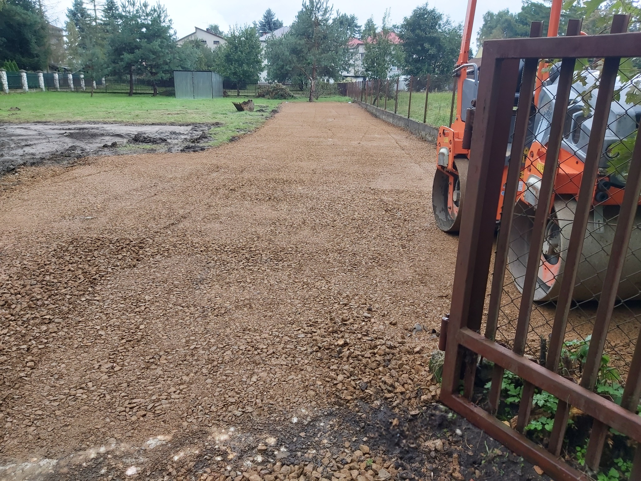 Pomarańczowy walec wibracyjny stoi obok utwardzonej, kamienistej drogi w trakcie budowy, widok przez otwartą, rdzawą furtkę.