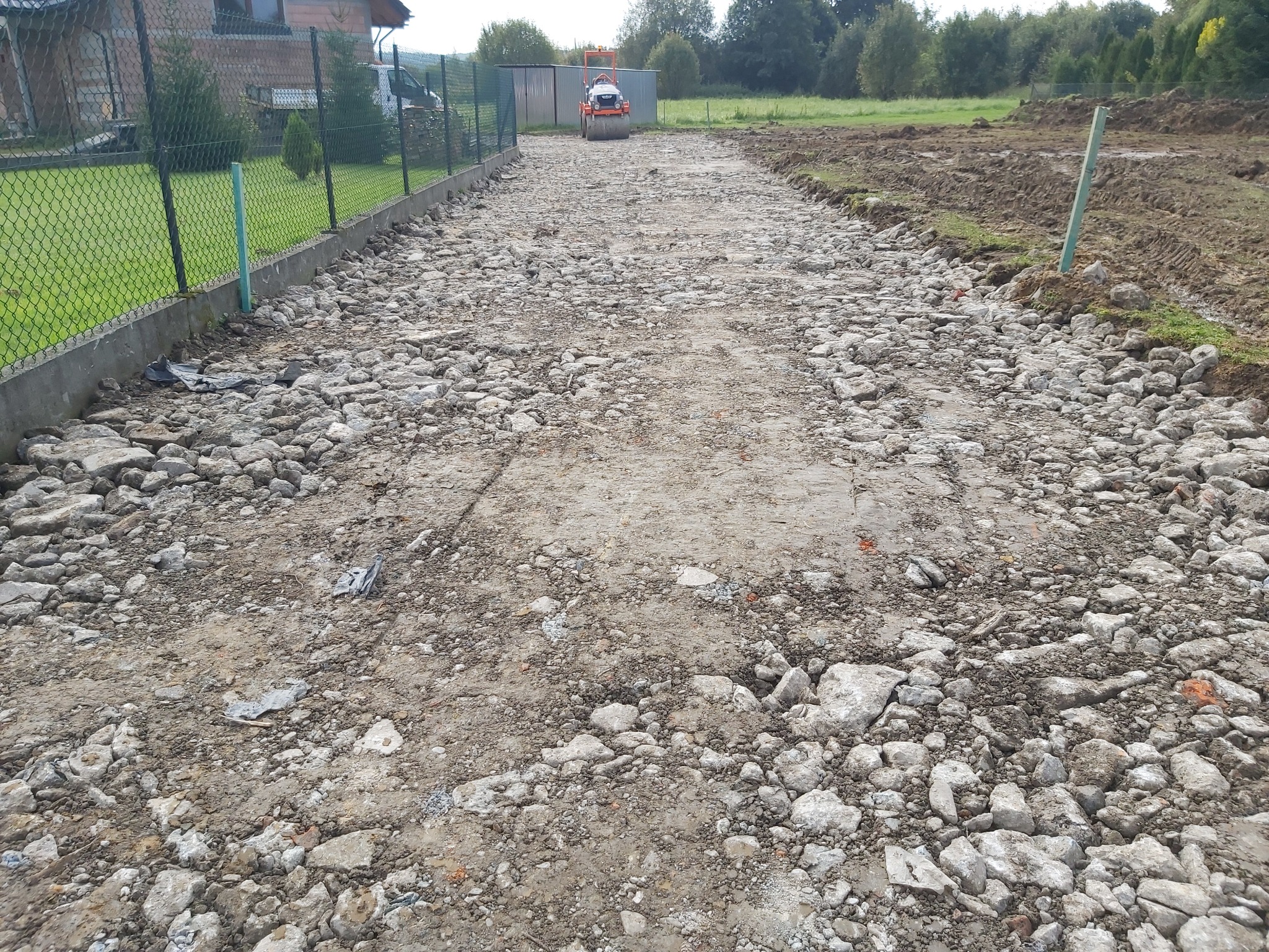 Budowa drogi dojazdowej: kruszywo betonowe zagęszczane walcem wibracyjnym, widoczna granica działki z ogrodzeniem.