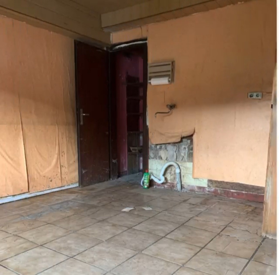 Pomieszczenie w trakcie remontu: uszkodzona ściana z odsłoniętym fragmentem muru, rury instalacyjne przy podłodze, stara terakota, drzwi wejściowe i fragment zakrytej ściany folią malarską.