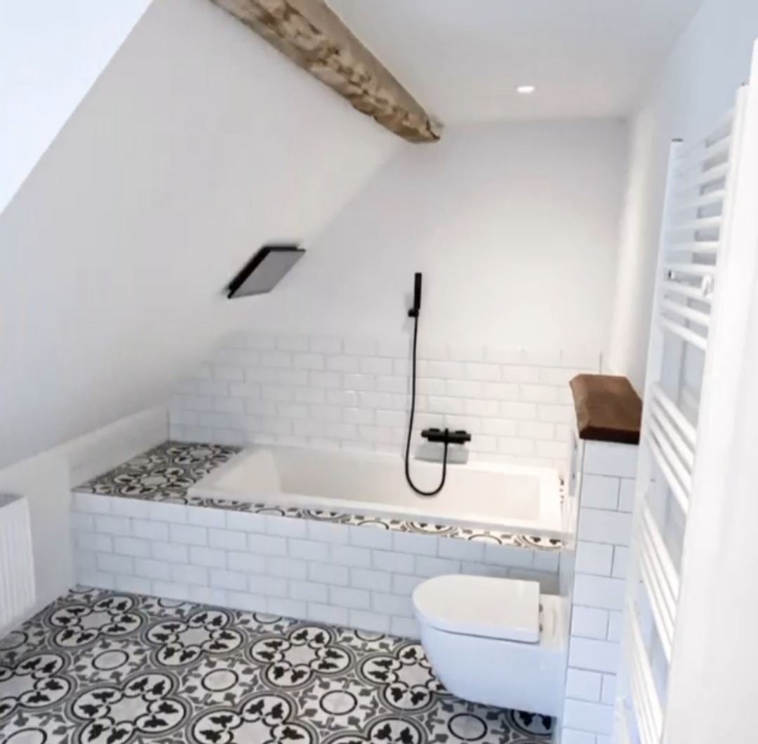 Biała łazienka na poddaszu z wanną obudowaną białymi płytkami, wzorzystą podłogą, czarną armaturą i drewnianą belką stropową