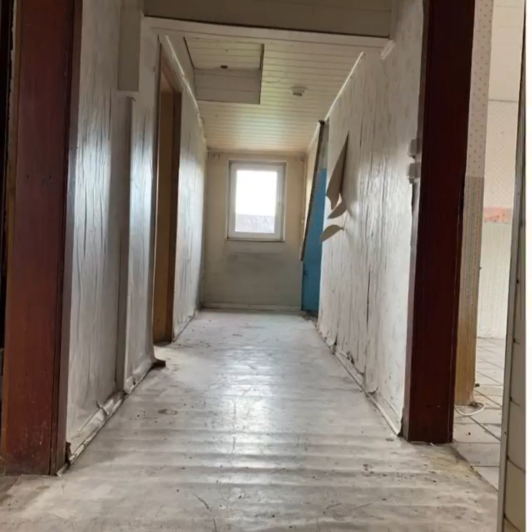 Korytarz w trakcie remontu: ściany częściowo pokryte szpachlą, widoczne odchodzące fragmenty starej tapety, surowa podłoga, okno na końcu korytarza.