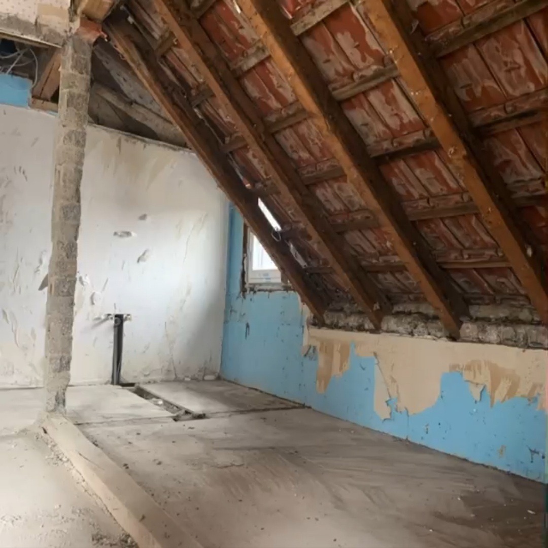 Poddasze w trakcie remontu: odsłonięta więźba dachowa z czerwonej dachówki, fragment ściany z pozostałościami niebieskiej farby, przygotowane podłoże pod posadzkę, widoczne instalacje.