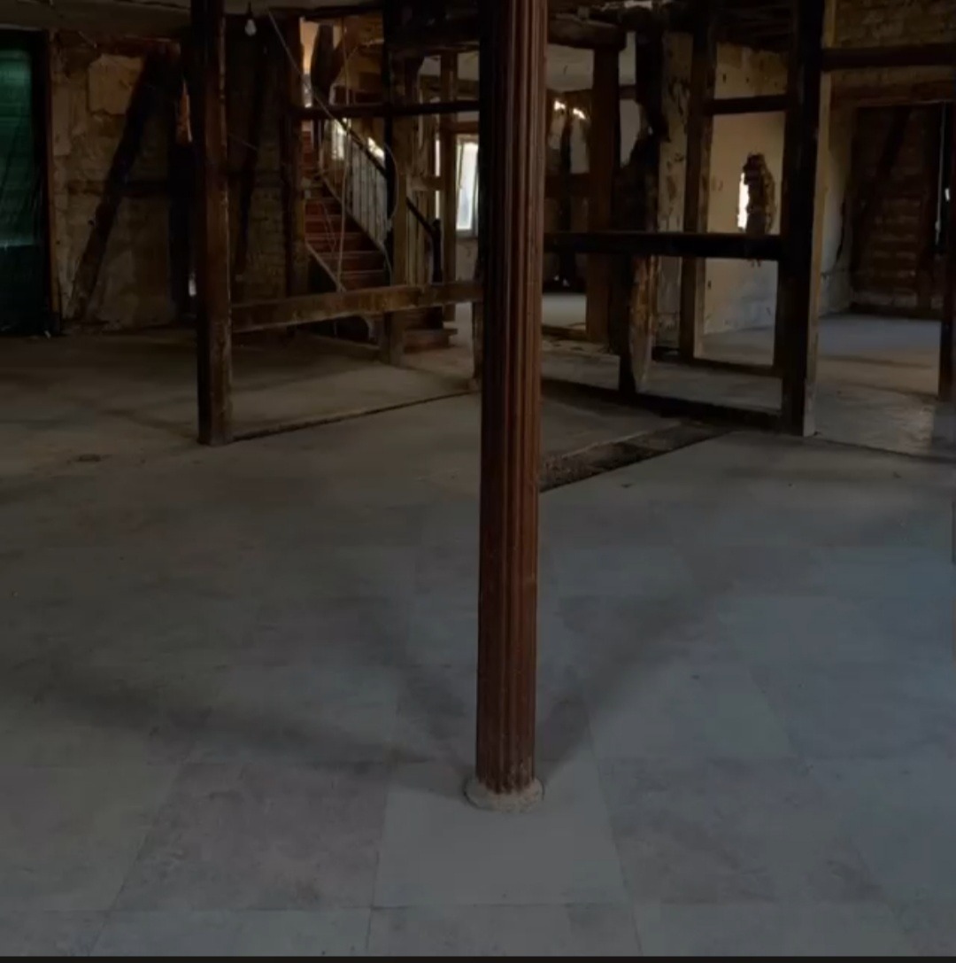 Wnętrze budynku w trakcie renowacji z widoczną drewnianą konstrukcją szkieletową, kolumną z dekoracyjnymi żłobieniami i marmurową podłogą w odcieniach szarości.