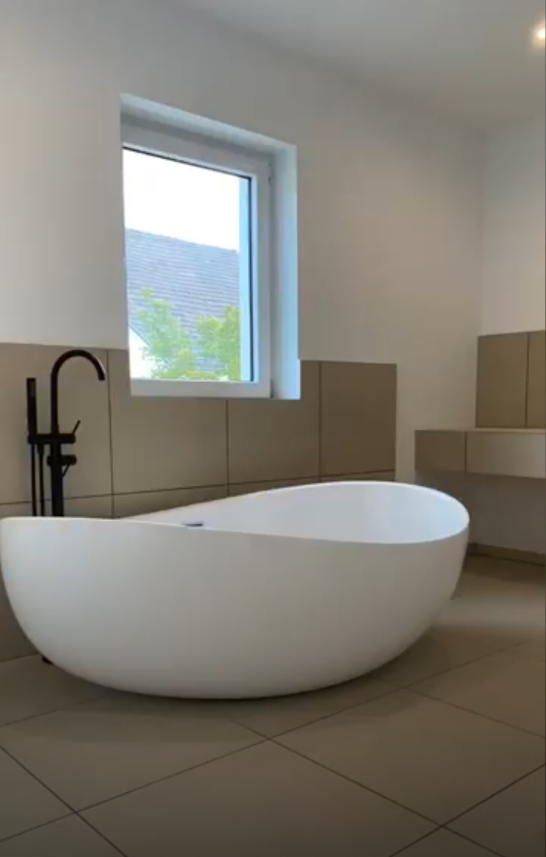 Nowoczesna, wolnostojąca wanna o asymetrycznym kształcie w minimalistycznej łazience z beżowymi płytkami i czarną armaturą, widok na okno z zielenią na zewnątrz.