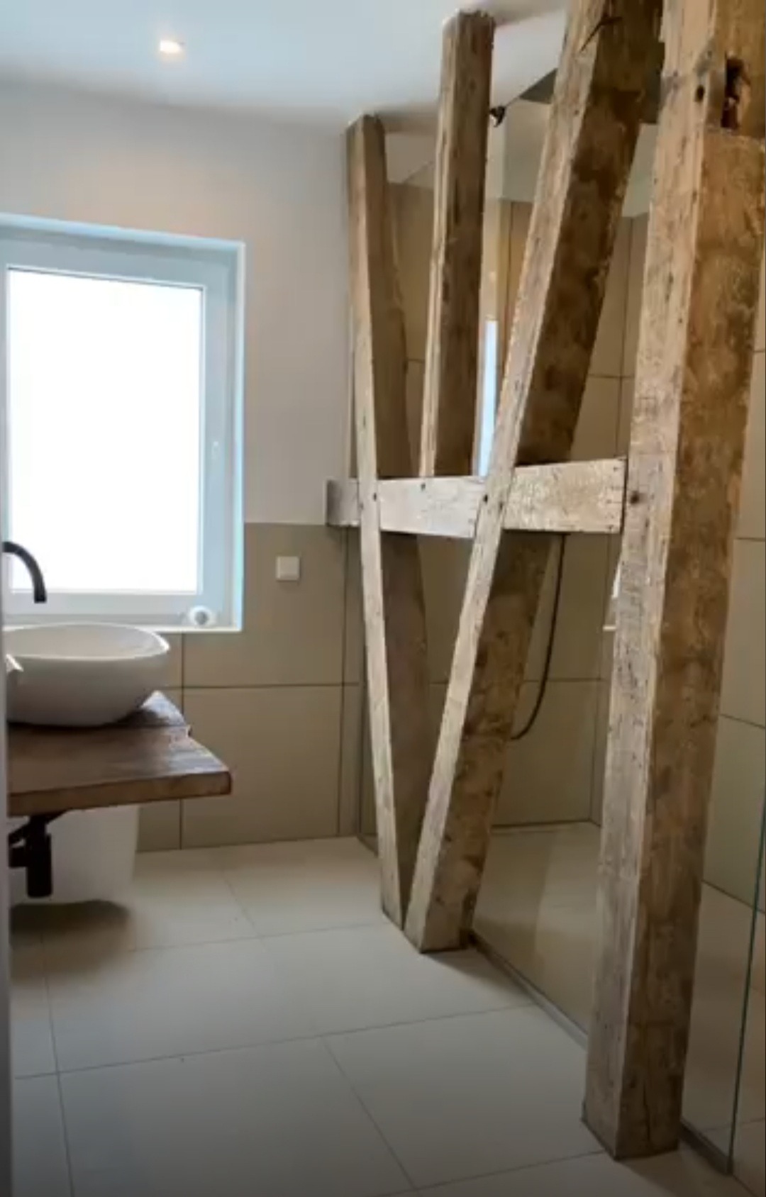 Nowoczesna łazienka z umywalką na drewnianym blacie i kabiną prysznicową oddzieloną drewnianymi belkami konstrukcyjnymi.