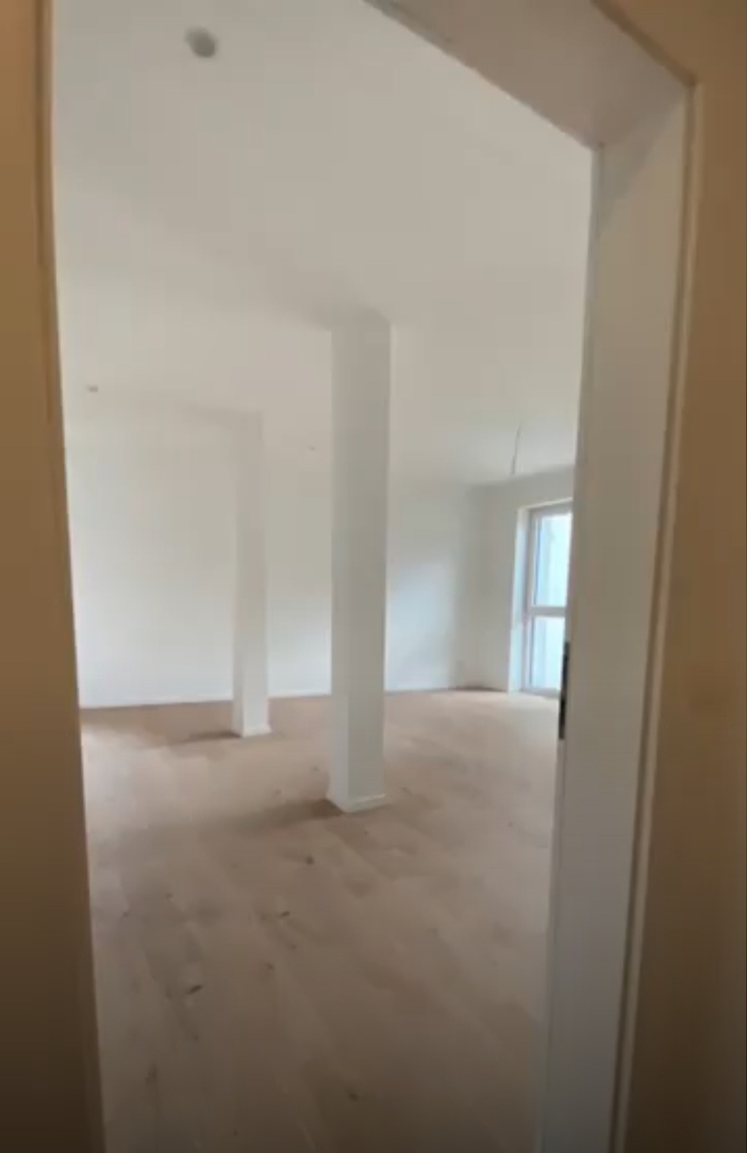 Jasne, przestronne wnętrze z drewnianą podłogą i białymi ścianami, widoczne przez framugę drzwi. Dwa białe filary podtrzymują sufit. Naturalne światło wpada przez okno w głębi pomieszczenia.