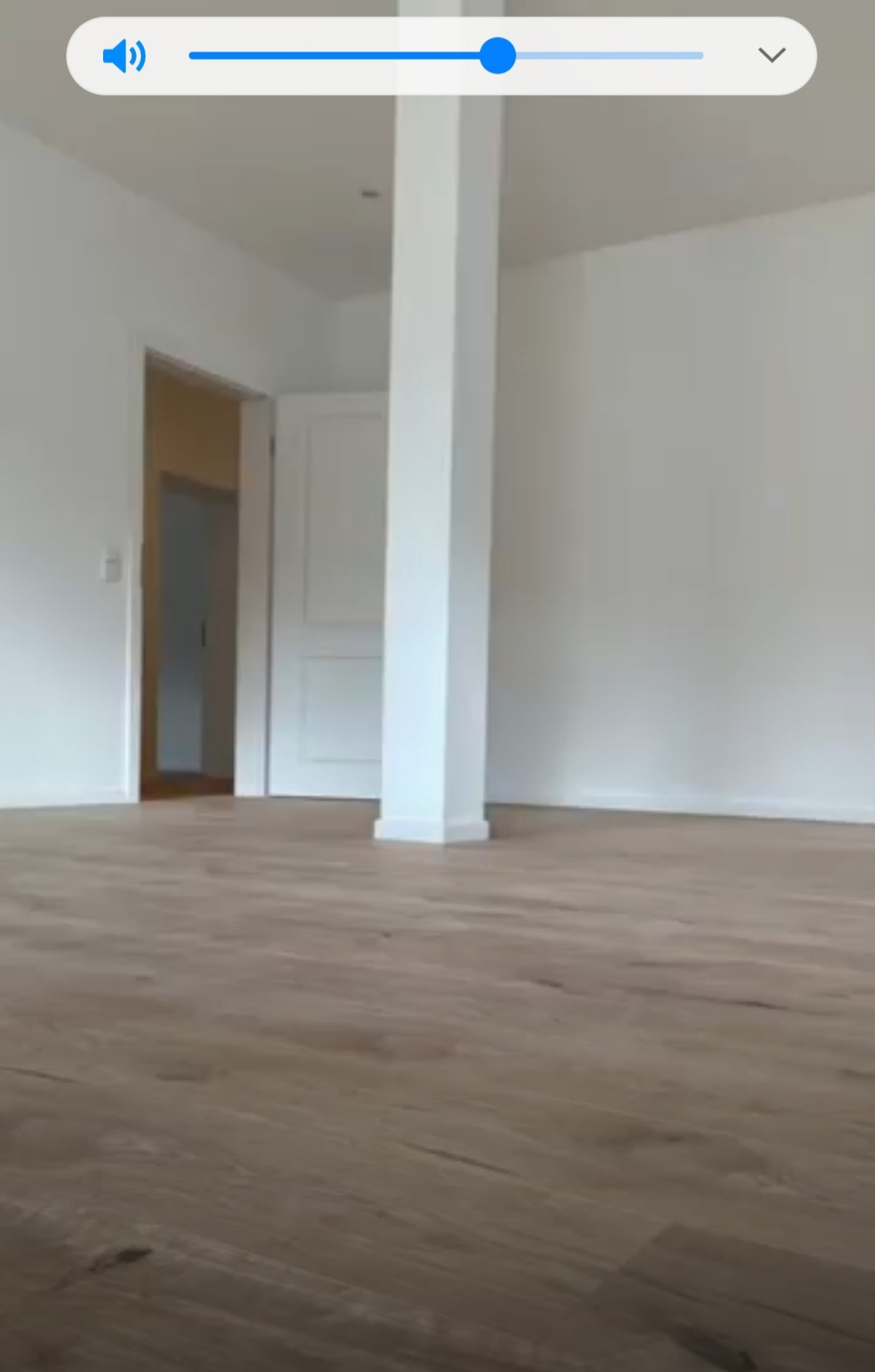 Pusta, biała przestrzeń z drewnianą podłogą, kolumną nośną i otwartymi drzwiami do kolejnego pomieszczenia, widok z poziomu podłogi.