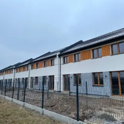 Szereg nowo wybudowanych segmentów mieszkalnych z drewnianą elewacją, ogrodzonych siatką na betonowej podmurówce, w pochmurny dzień.