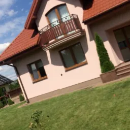 Ocieplony dom jednorodzinny z beżową elewacją, brązową stolarką okienną i balkonem z kutą balustradą, widok z poziomu trawnika.