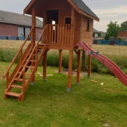 Drewniany domek dla dzieci na podwyższeniu z czerwoną zjeżdżalnią, umieszczony na trawniku przed zabudowaniami gospodarczymi.