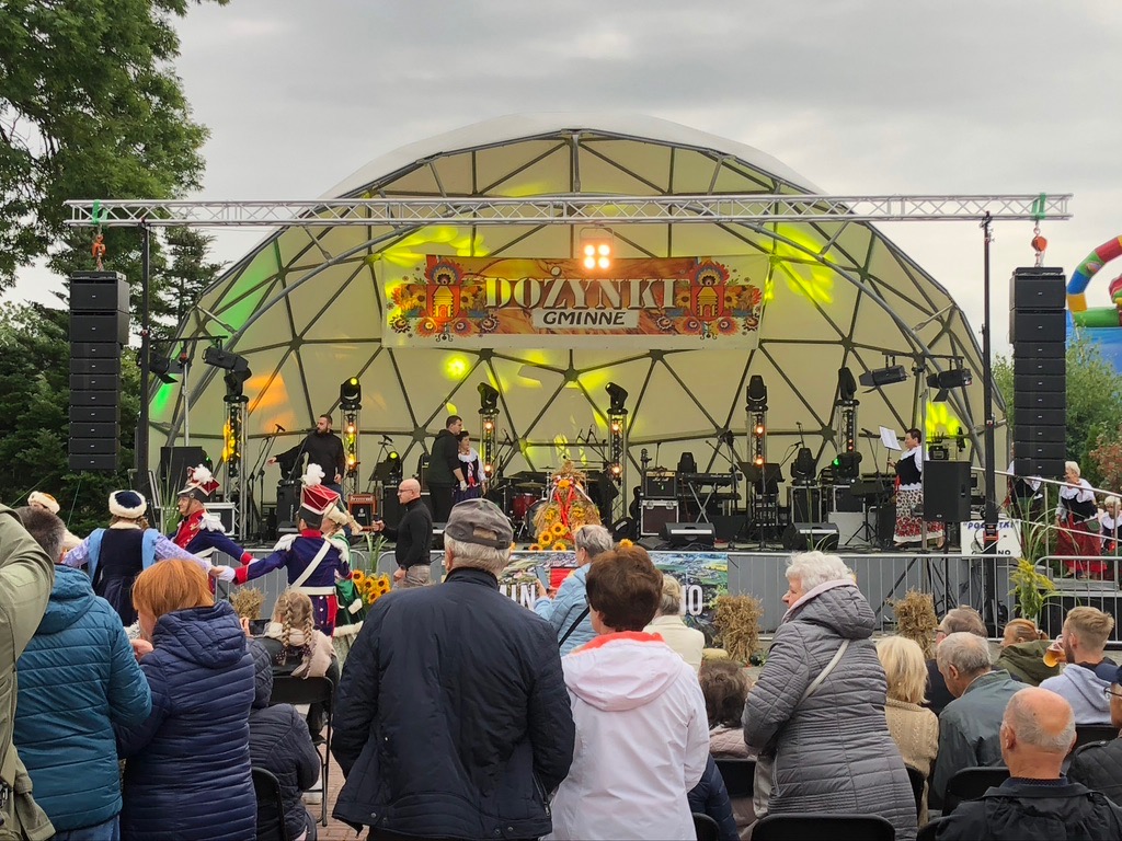 Scena plenerowa z zespołem muzycznym i tancerzami w strojach ludowych podczas Dożynek Gminnych, oświetlona reflektorami, z widownią na pierwszym planie.