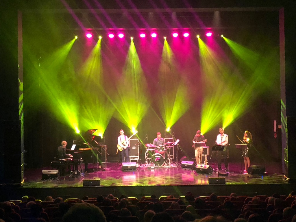 Zespół muzyczny na scenie oświetlonej zielonymi i różowymi reflektorami, widownia w tle.
