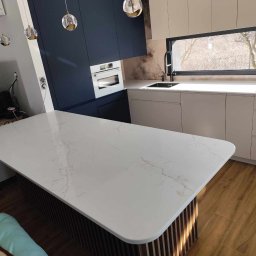 Fado Wood - Nowoczesna kuchnia z wyspą o marmurowym blacie, ciemnoniebieskie szafki górne i jasne dolne. Okno z widokiem na drzewa. Oświetlenie punktowe nad wyspą.