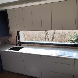 Fado Wood - Nowoczesna kuchnia z oknem: białe szafki dolne i górne, czarny zlew i bateria, marmurowy blat, zabudowany piekarnik. Widok na drzewa za oknem.