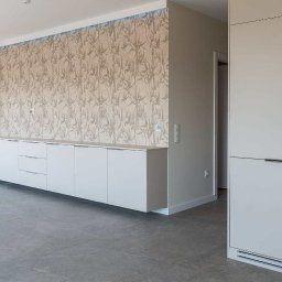 Fado Wood - Nowoczesna zabudowa meblowa w salonie z tapetą w roślinny wzór. Białe szafki i szuflady pod ścianą, obok zabudowa kuchenna z piekarnikiem. Szare płytki na podłodze.