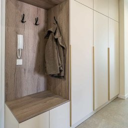 Fado Wood - Biała szafa wnękowa z drewnianym panelem, wieszakami i interkomem w przedpokoju. Minimalistyczny design, złote uchwyty, płaszcz na wieszaku.