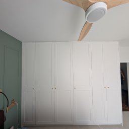 Fado Wood - Biała szafa wnękowa z sześcioma drzwiami, złotymi gałkami, w sypialni z zieloną ścianą i drewnianym wentylatorem sufitowym. Widok na otwarte drzwi do innego pokoju.