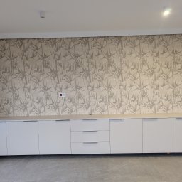 Fado Wood - Długa, biała szafka pod ścianą z tapetą w motywy roślinne. Minimalistyczny design, brak widocznych uchwytów. Szara podłoga. Nowoczesne wnętrze.