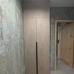 Fado Wood - Jasna szafa z drewnianymi frontami i długimi, czarnymi uchwytami, na tle ściany z dekoracyjnym tynkiem i otwartych, drewnianych drzwi.