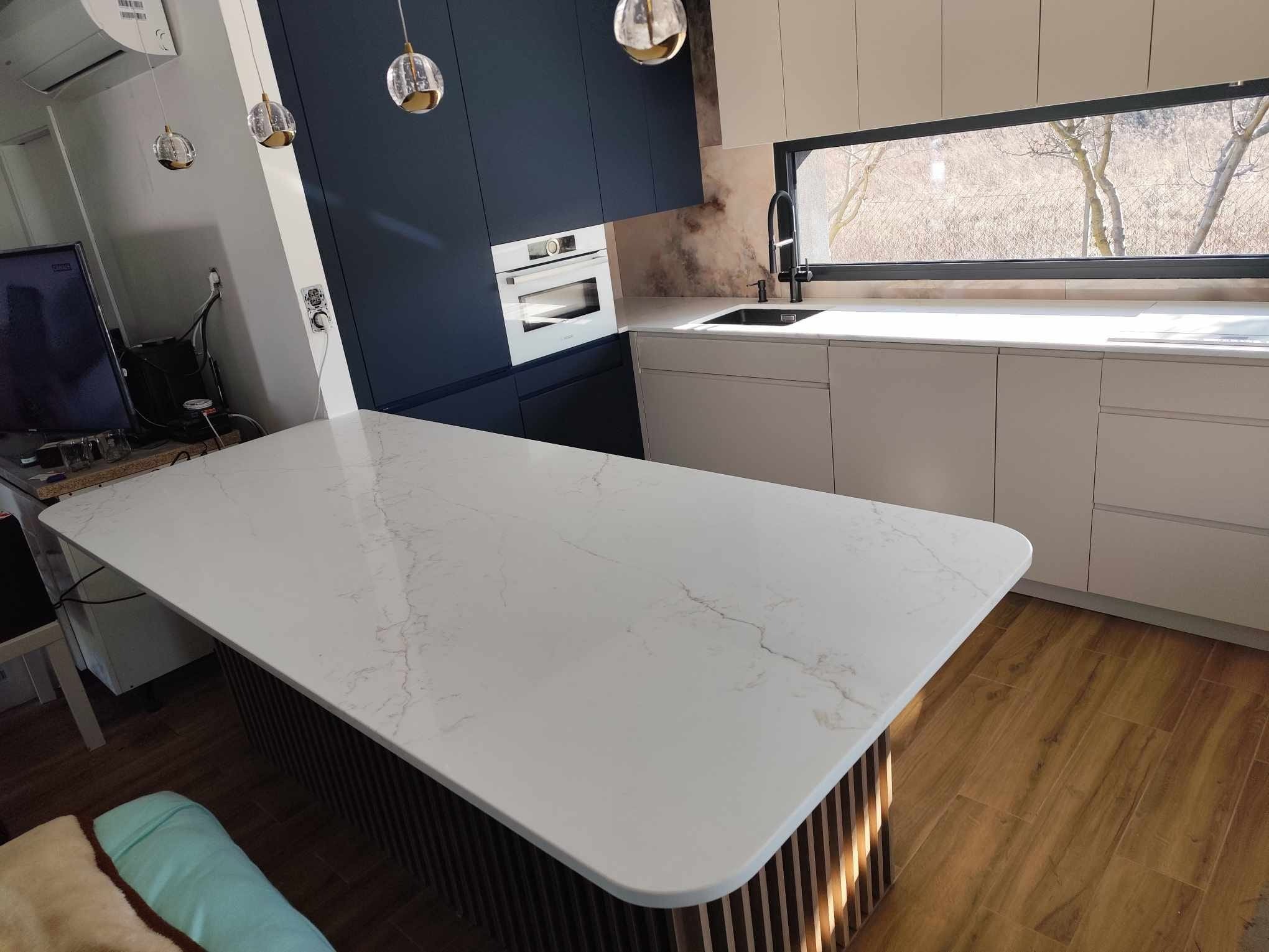 Nowoczesna kuchnia z wyspą o marmurowym blacie, ciemnoniebieskie szafki górne i jasne dolne. Okno z widokiem na drzewa. Oświetlenie punktowe nad wyspą.