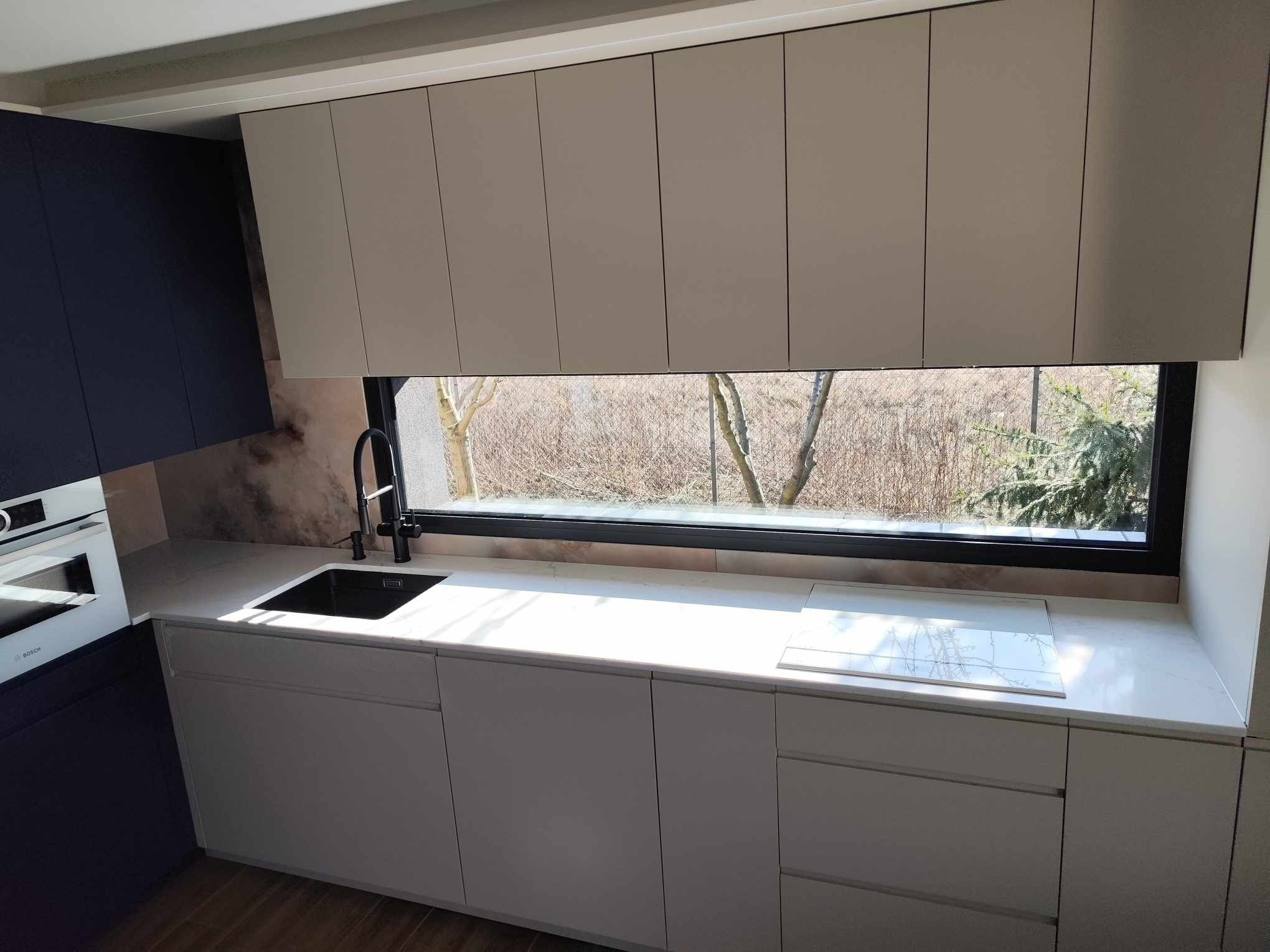 Nowoczesna kuchnia z oknem: białe szafki dolne i górne, czarny zlew i bateria, marmurowy blat, zabudowany piekarnik. Widok na drzewa za oknem.