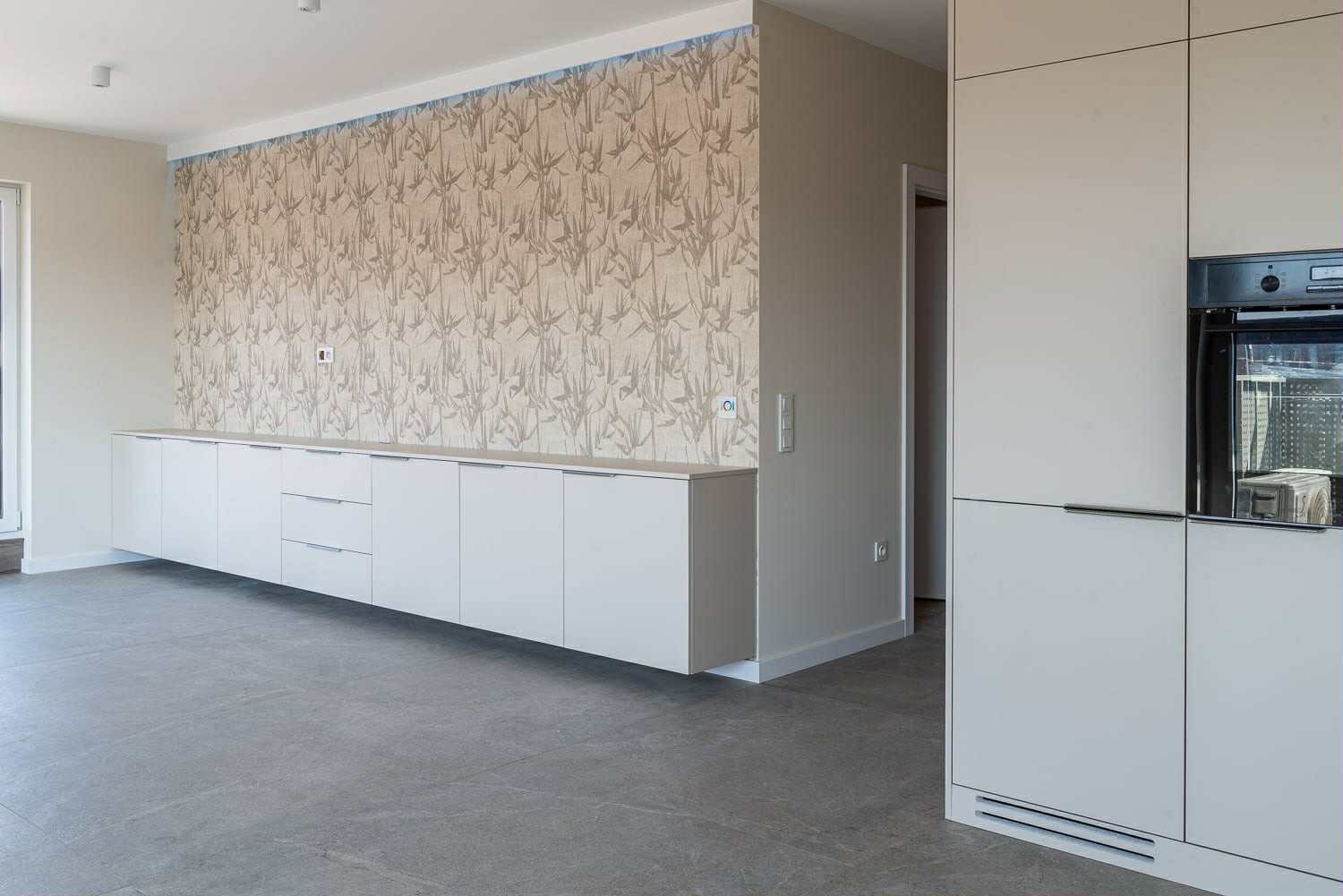 Nowoczesna zabudowa meblowa w salonie z tapetą w roślinny wzór. Białe szafki i szuflady pod ścianą, obok zabudowa kuchenna z piekarnikiem. Szare płytki na podłodze.