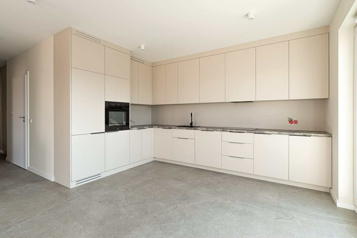 Jasna, narożna kuchnia z zabudową pod sufit, szarym blatem i płytkami podłogowymi. Minimalistyczny design, czarna armatura i sprzęt AGD. Widok na całe pomieszczenie.