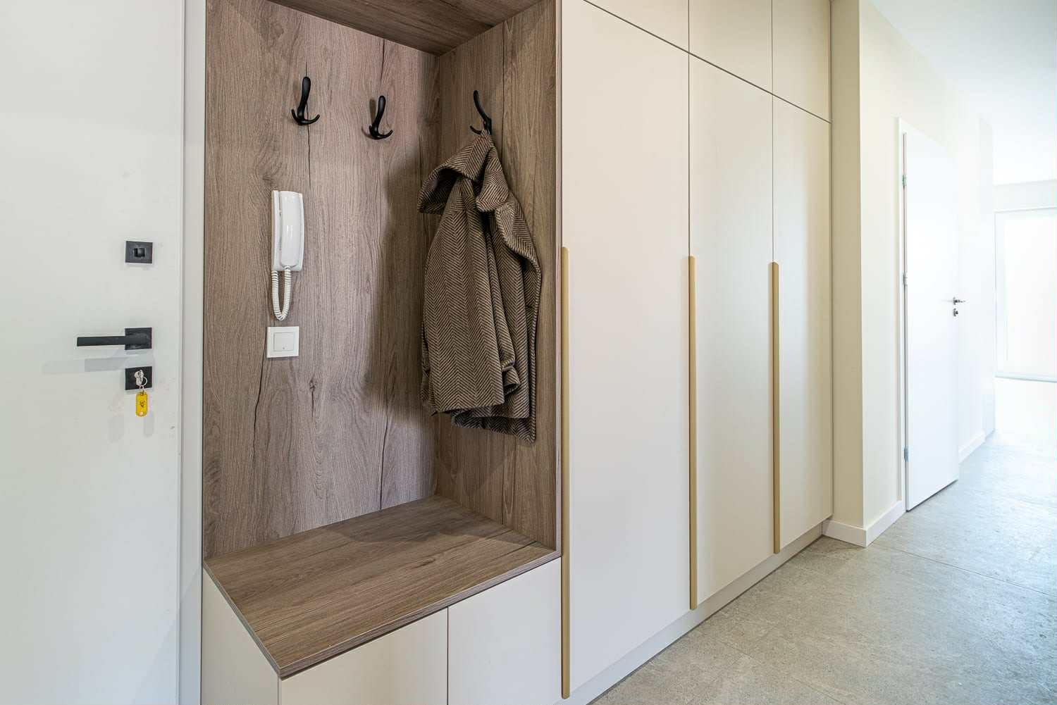 Biała szafa wnękowa z drewnianym panelem, wieszakami i interkomem w przedpokoju. Minimalistyczny design, złote uchwyty, płaszcz na wieszaku.