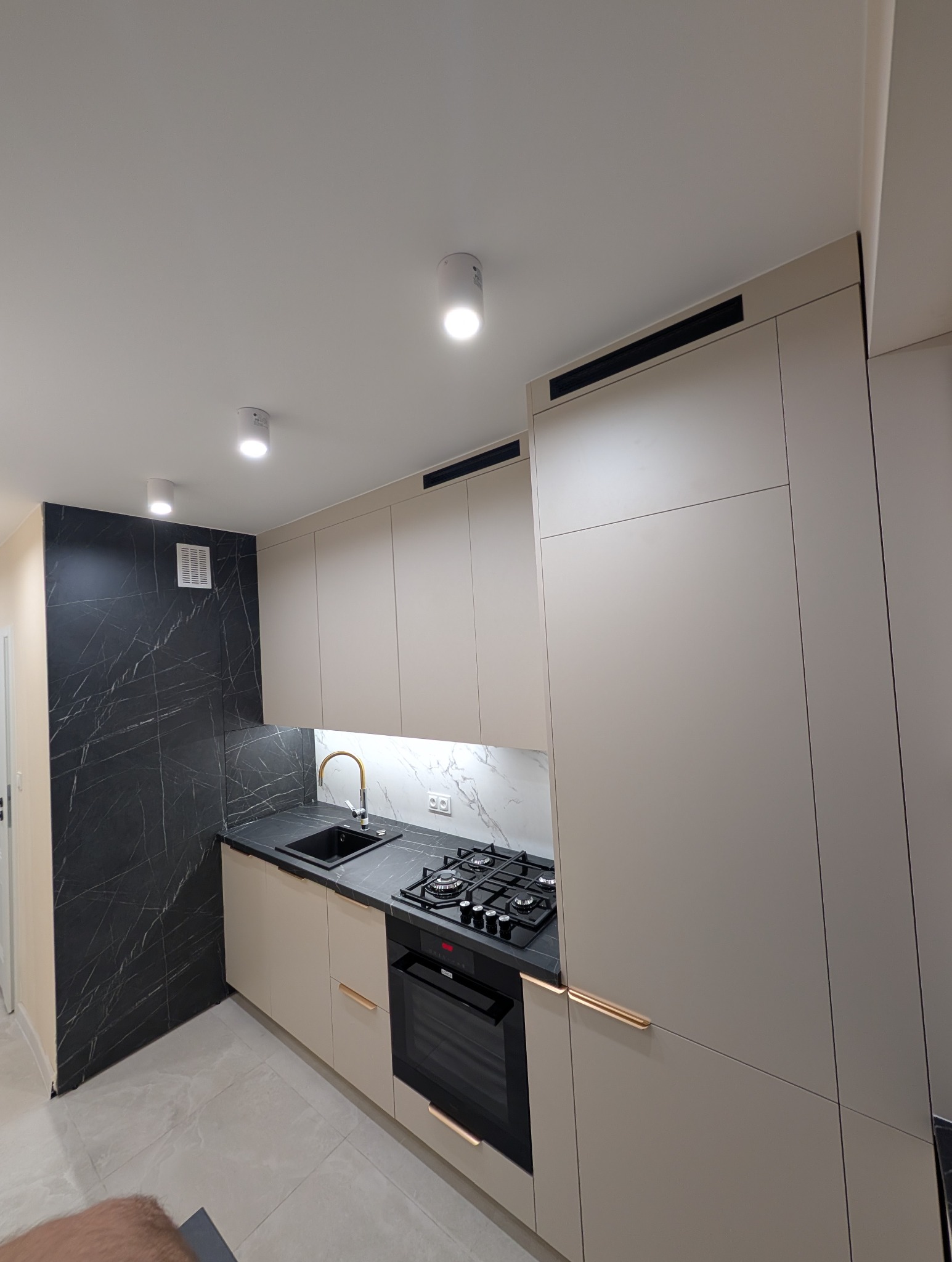 Nowoczesna kuchnia z beżowymi szafkami, czarnym blatem i marmurowym backsplashiem. Złote uchwyty dodają elegancji. Oświetlenie punktowe podkreśla detale.