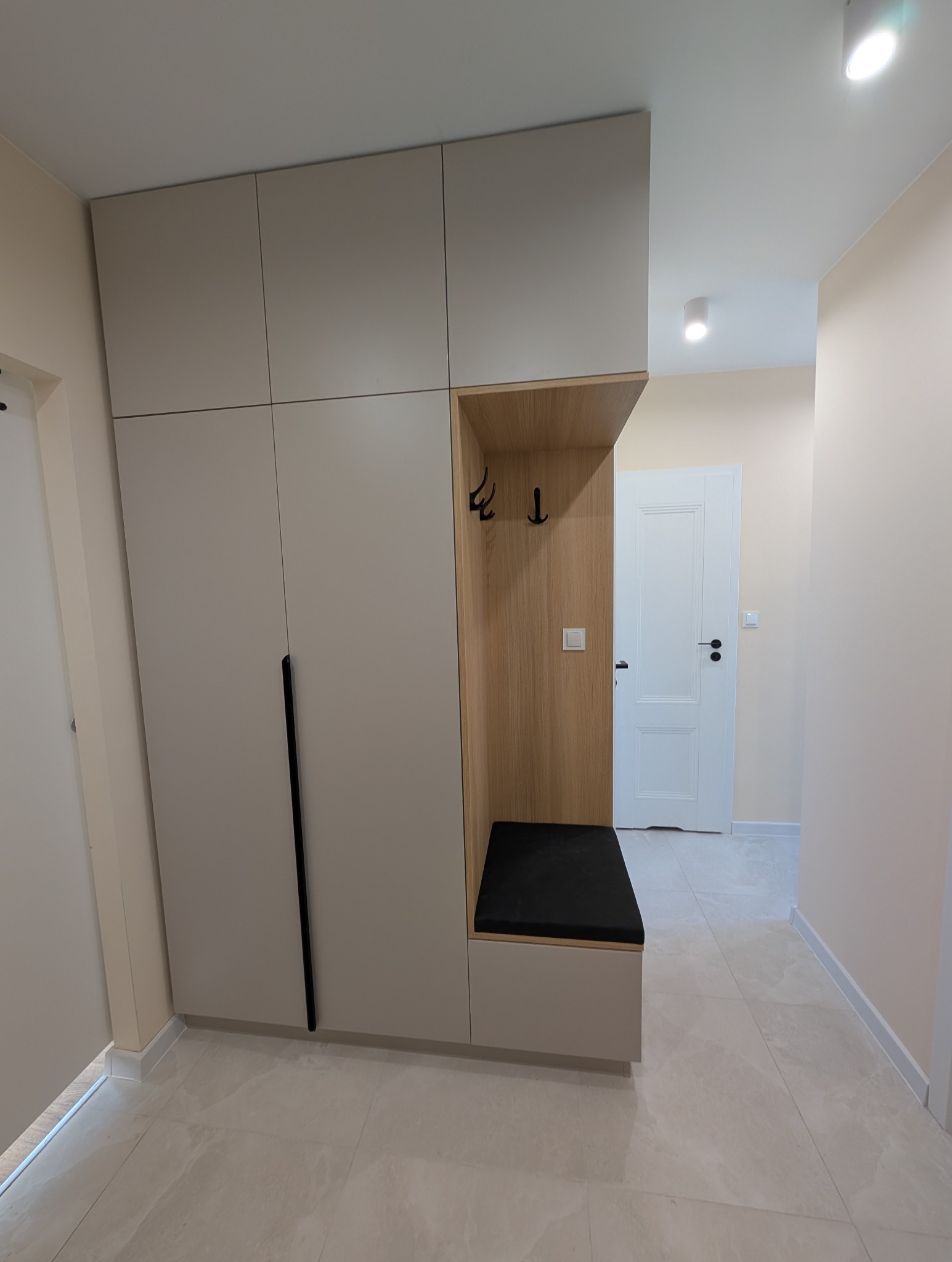 Jasna szafa wnękowa z siedziskiem i wieszakami w przedpokoju. Minimalistyczny design, gładkie fronty, czarne uchwyty. Drewniane wykończenie wnęki.
