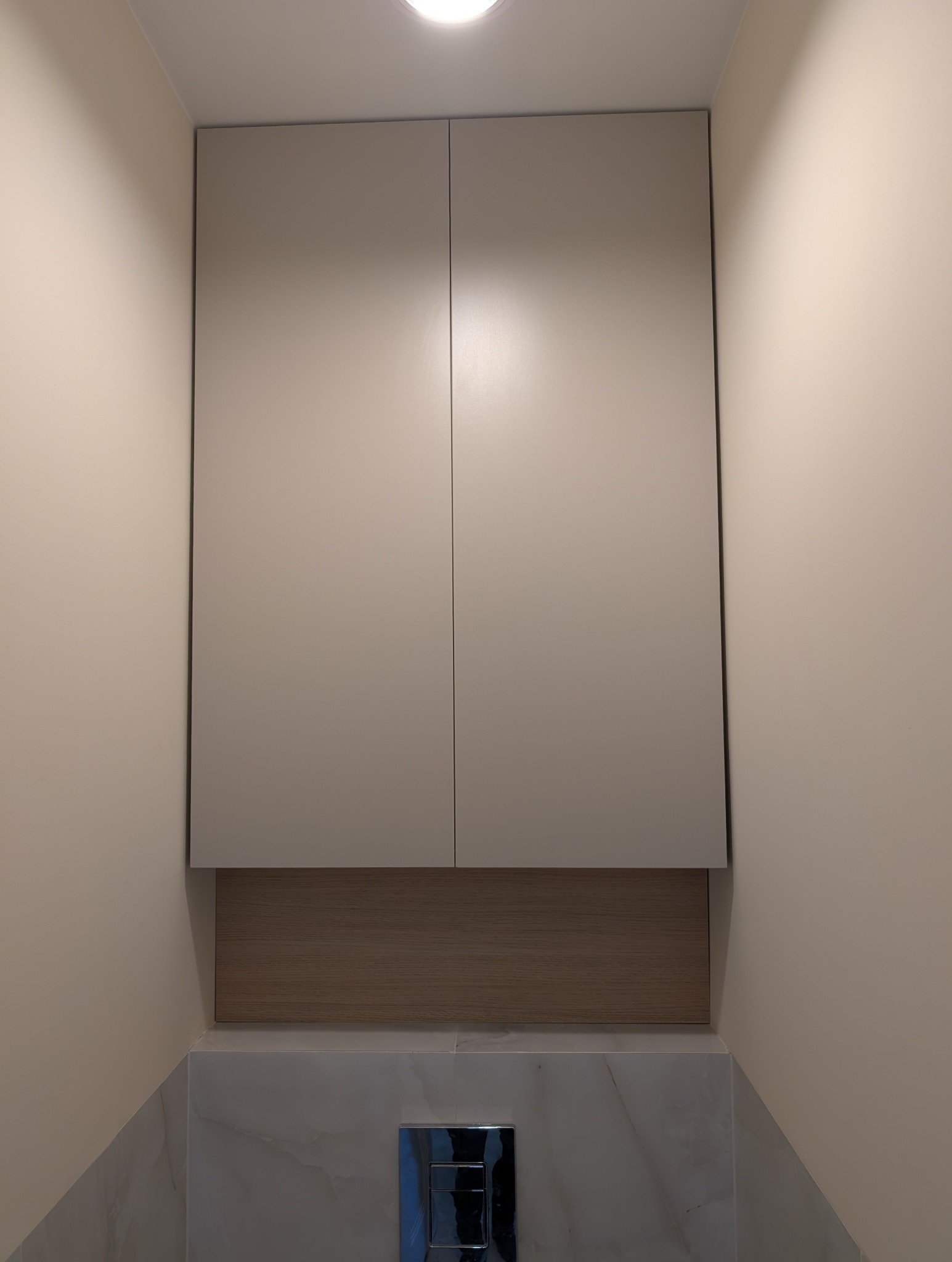 Zamknięta, beżowa szafka wisząca nad toaletą z drewnianym panelem i chromowanym przyciskiem spłukiwania na tle marmurowych płytek i beżowej ściany.