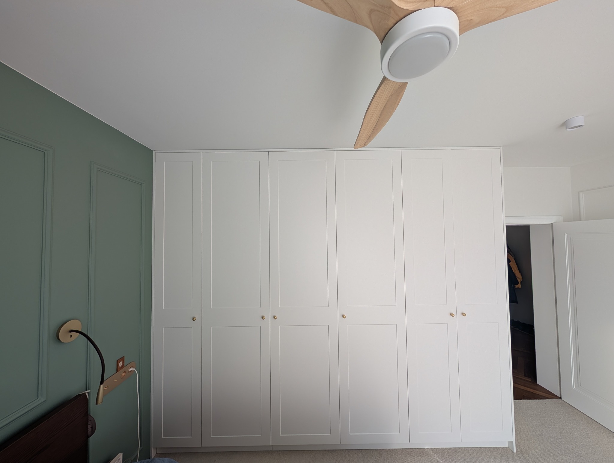 Biała szafa wnękowa z sześcioma drzwiami, złotymi gałkami, w sypialni z zieloną ścianą i drewnianym wentylatorem sufitowym. Widok na otwarte drzwi do innego pokoju.