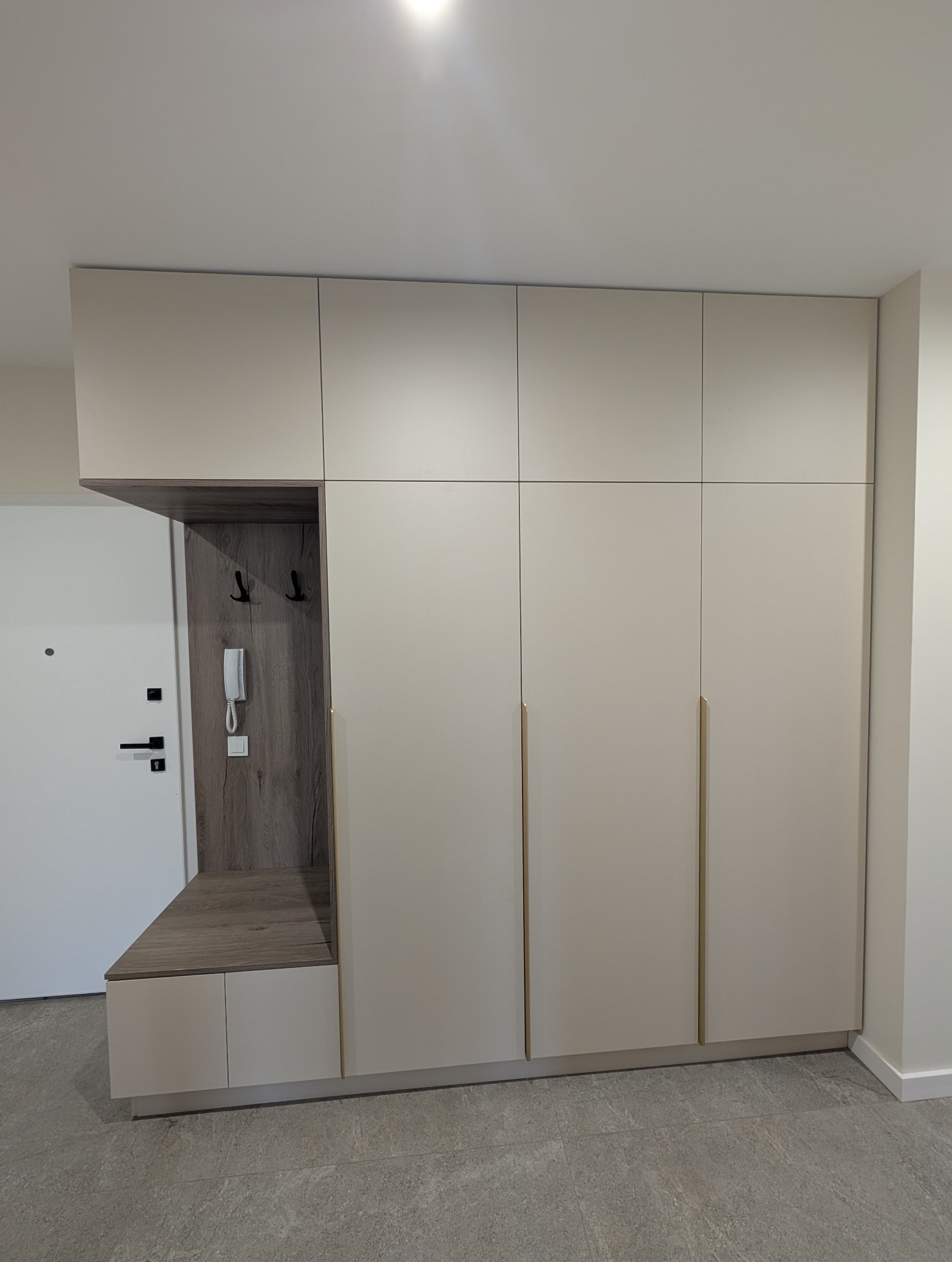 Beżowa szafa wnękowa z siedziskiem i wieszakami, zintegrowana z przedpokojem. Minimalistyczny design, złote uchwyty, drewnopodobne wykończenie wnęki.