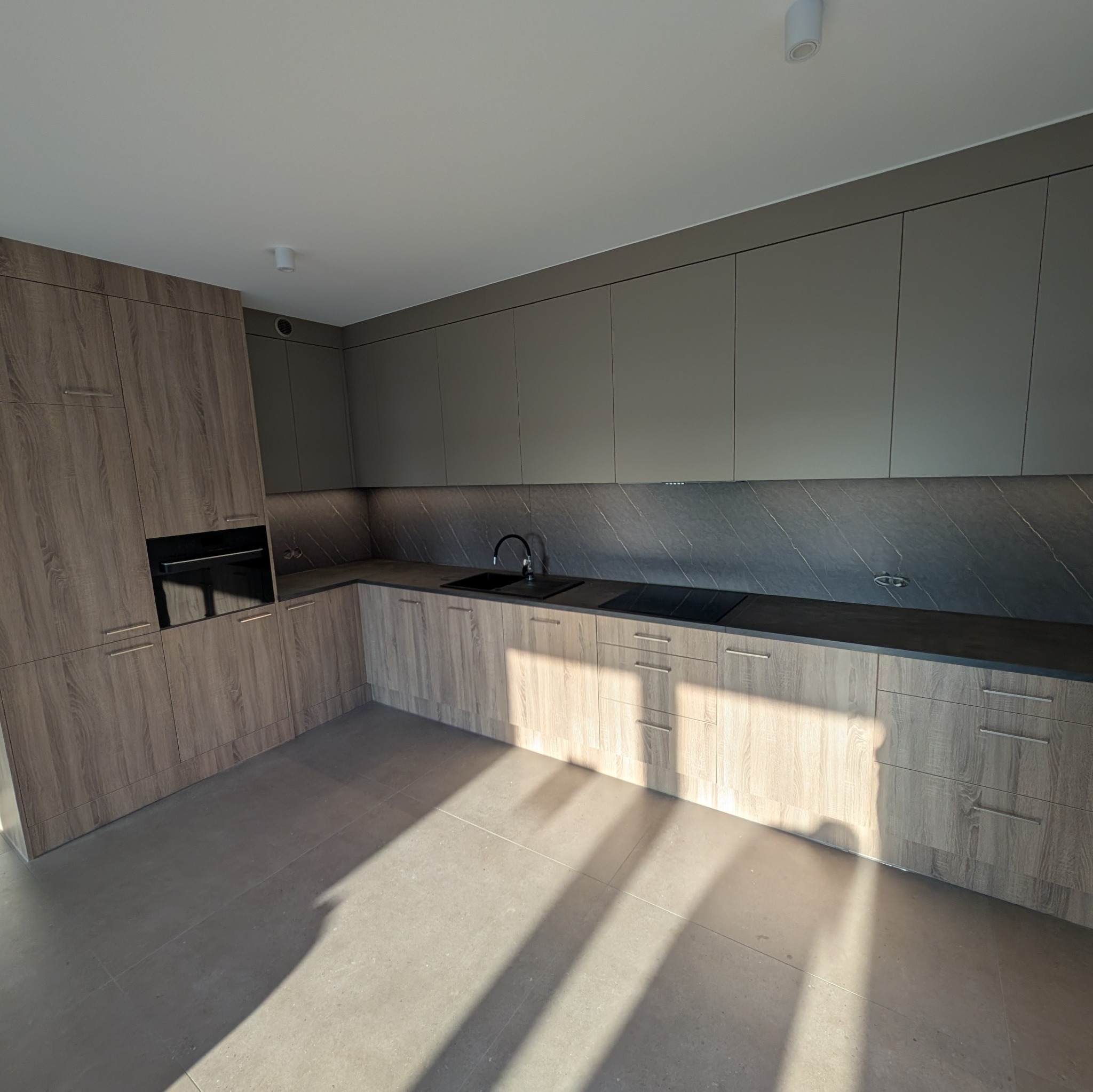 Nowoczesna kuchnia z drewnianymi i szarymi szafkami, czarnym blatem i marmurowym backsplashiem. Widoczne oświetlenie sufitowe i cień padający na podłogę.
