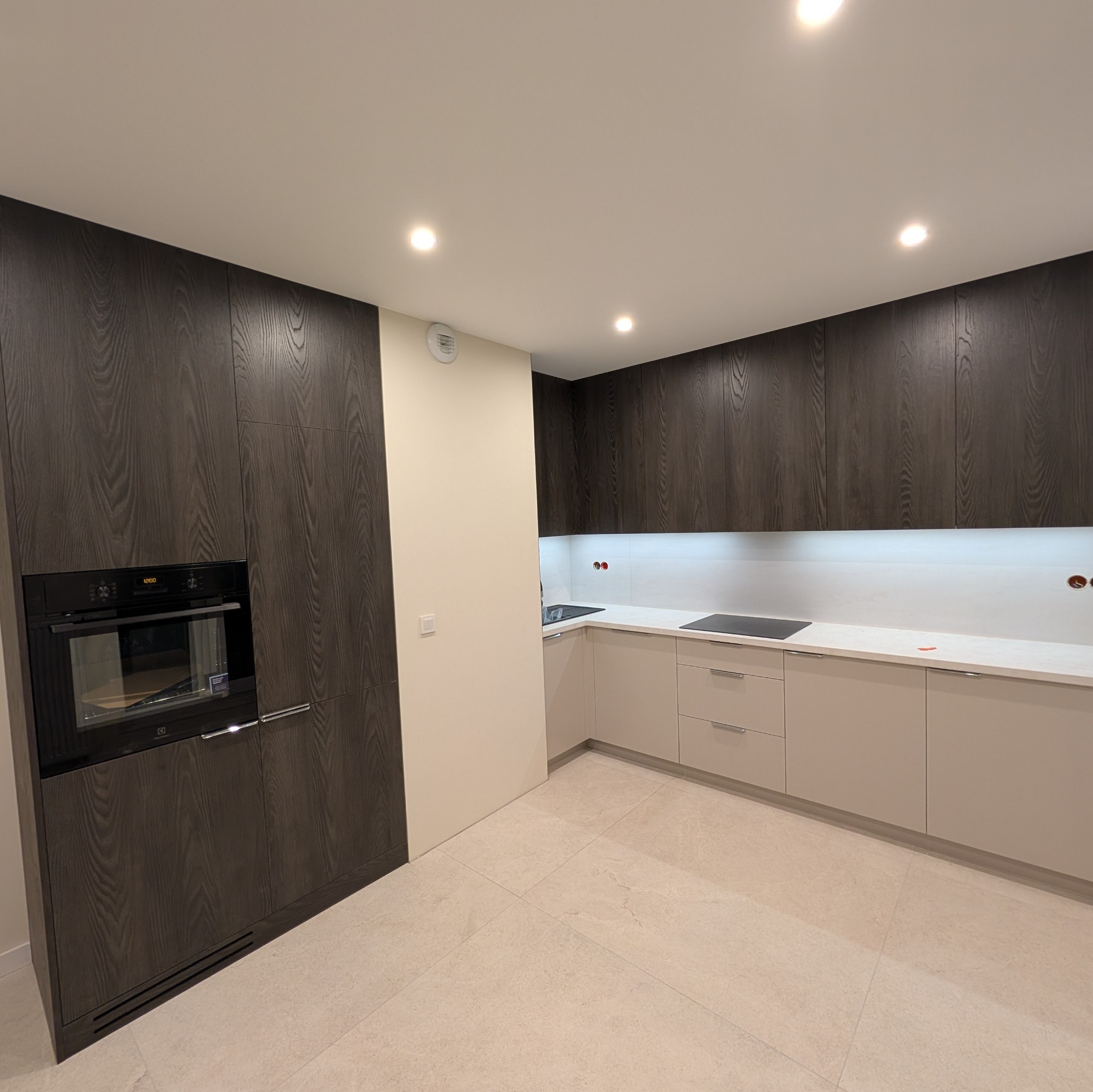 Nowoczesna kuchnia z ciemnymi, drewnianymi szafkami i beżowymi dolnymi szafkami. W zabudowie piekarnik. Blat biały, oświetlenie punktowe.