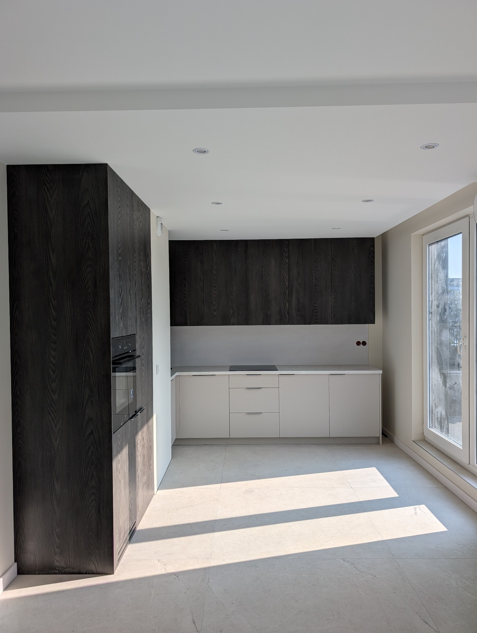 Nowoczesna kuchnia z ciemnymi, drewnianymi szafkami górnymi i jasnymi dolnymi, minimalistyczny design, oświetlenie punktowe, jasna podłoga i okno z widokiem na miasto.