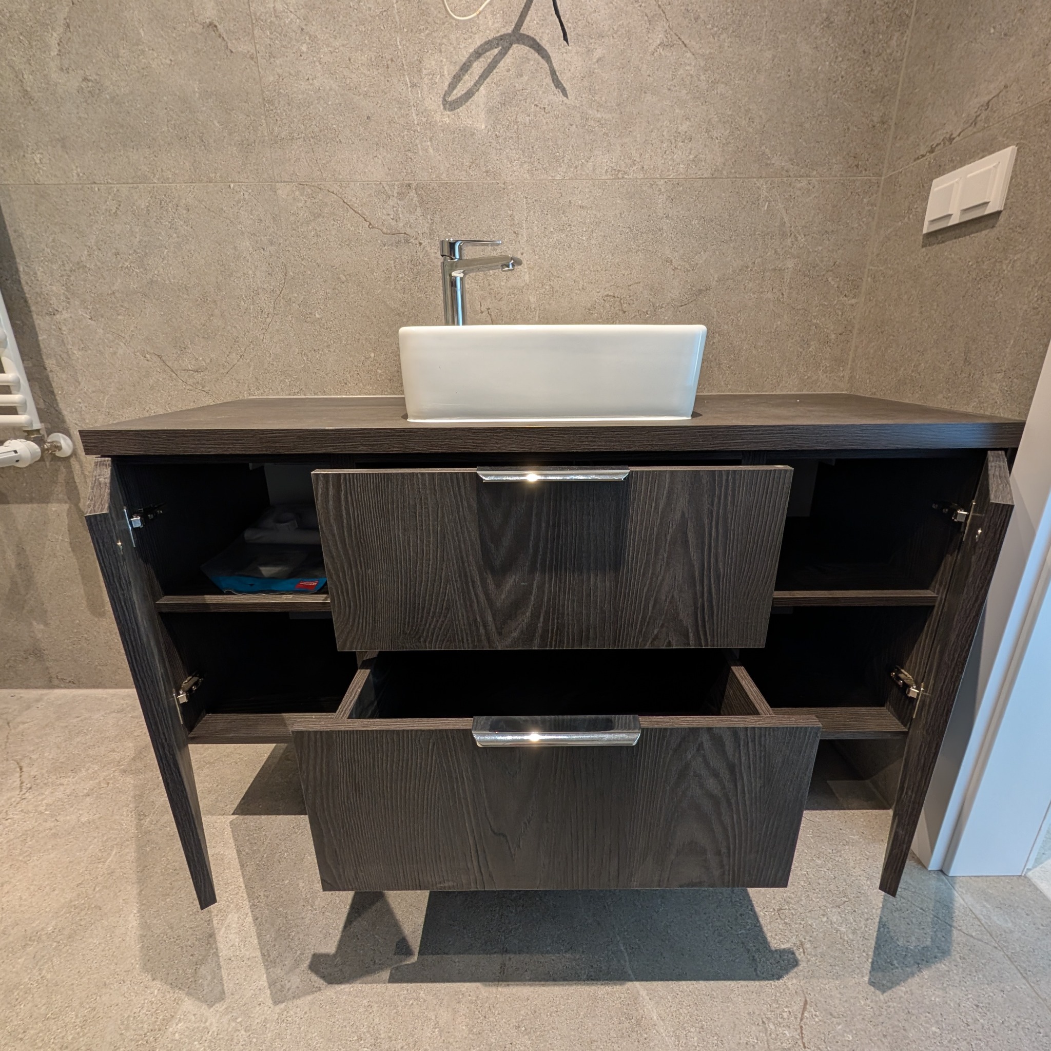 Ciemna szafka łazienkowa z dwoma szufladami i otwartymi drzwiczkami, na niej biała umywalka nablatowa i chromowana bateria. Szare płytki na ścianie i podłodze.