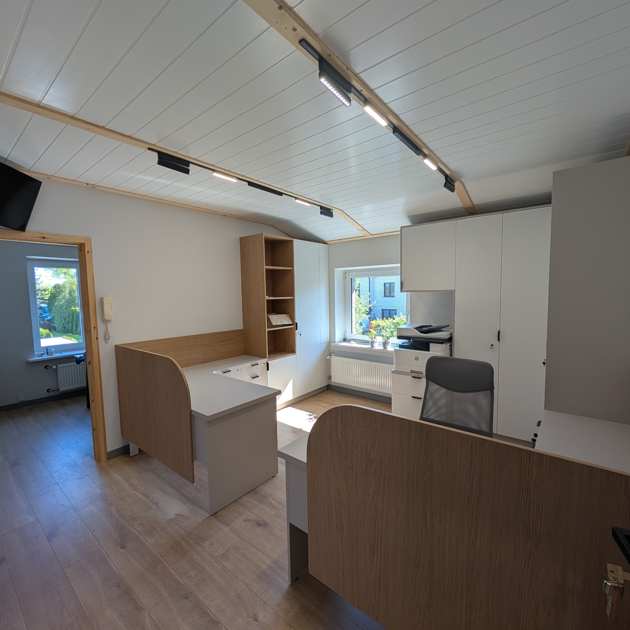 Jasne biuro z meblami na wymiar: biurka z drewnianymi panelami, białe szafy i regał. Widok na okno z roślinami i fragment pomieszczenia z drzwiami.