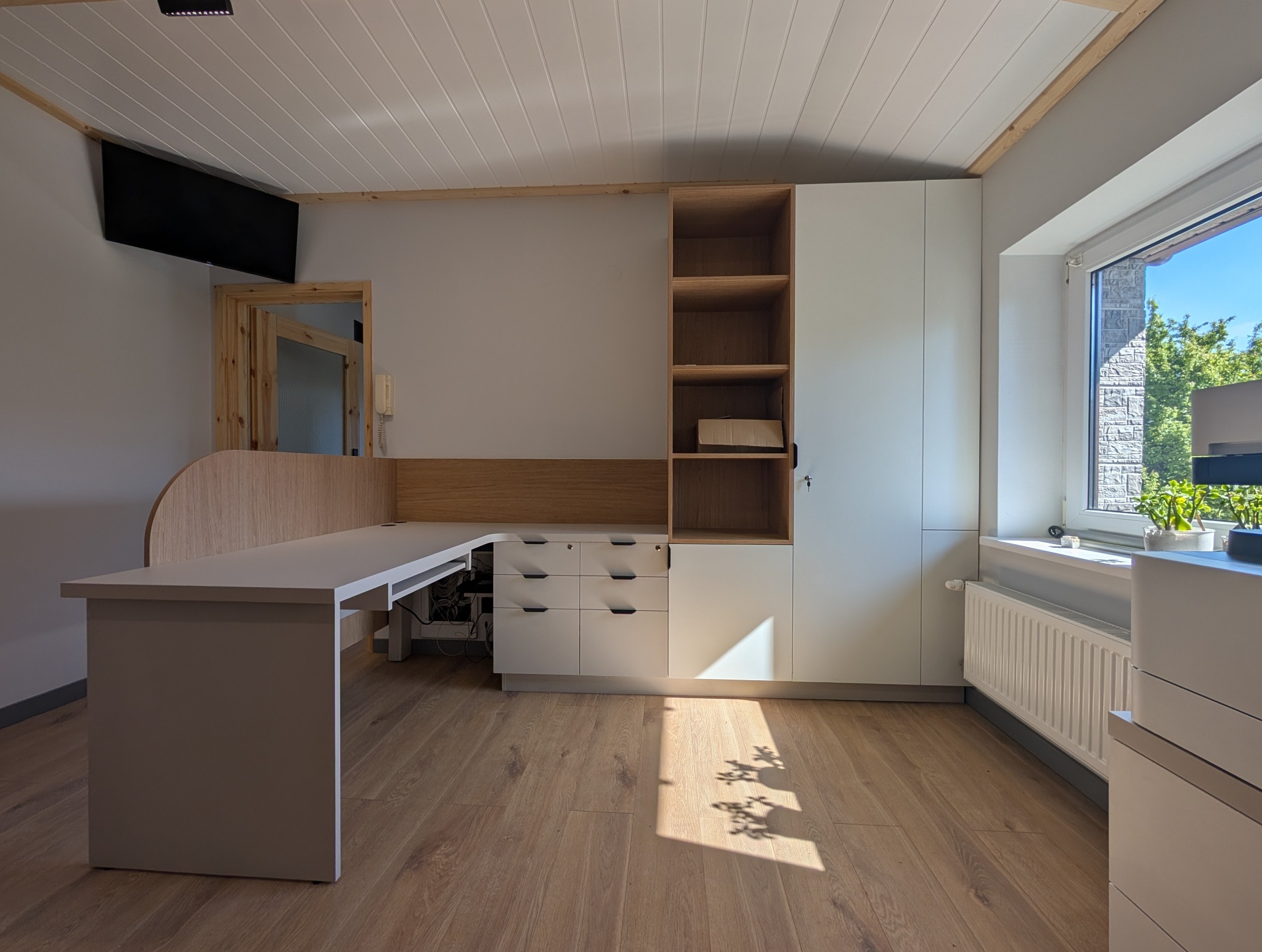 Nowoczesny pokój z biurkiem na wymiar, białe szafy z drewnianymi akcentami, drewniana podłoga i okno z widokiem na zieleń. Minimalistyczny design, jasne wnętrze.