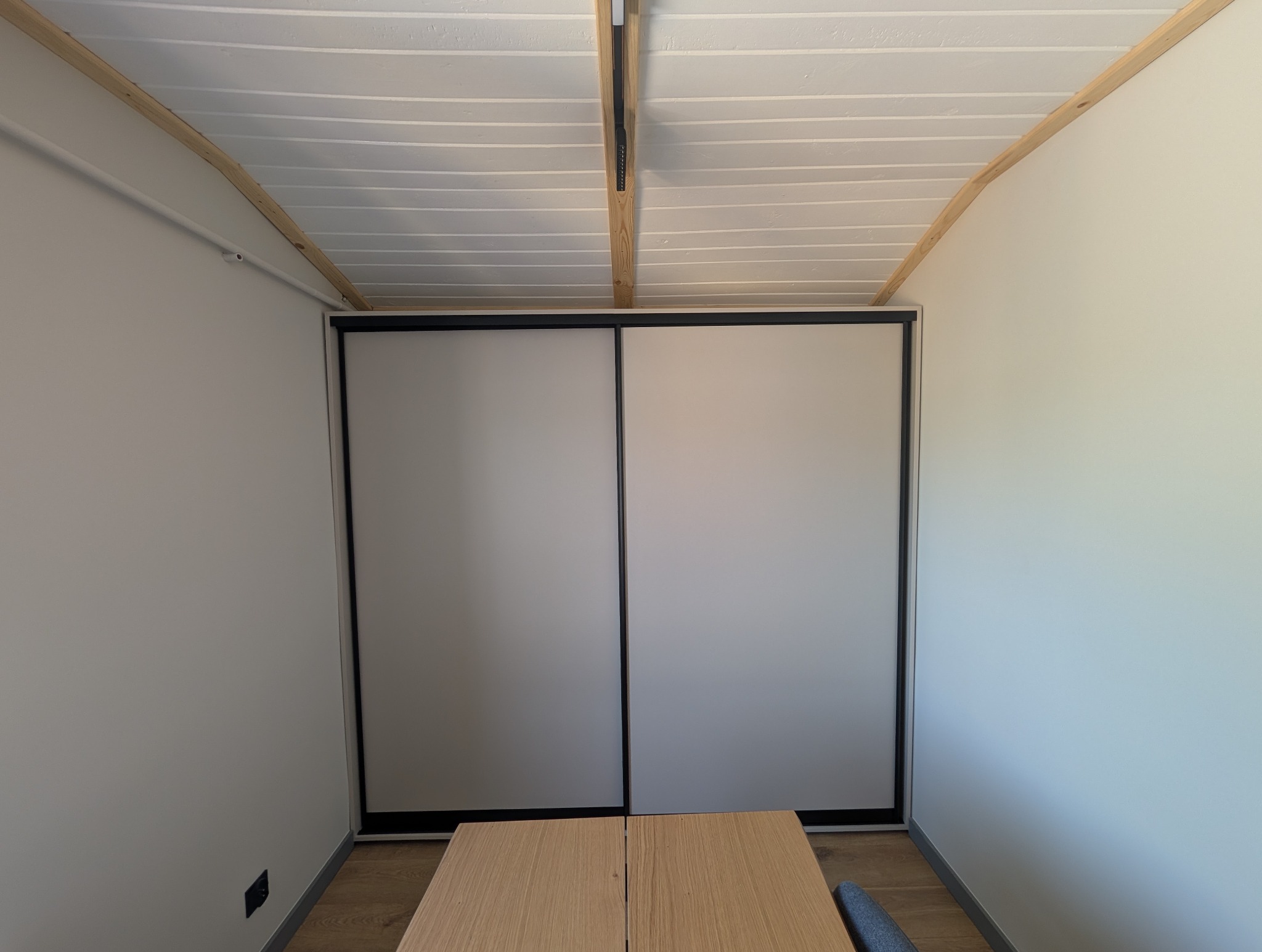 Minimalistyczna szafa wnękowa z przesuwnymi drzwiami w jasnym pomieszczeniu z drewnianym sufitem i fragmentem stołu na pierwszym planie.