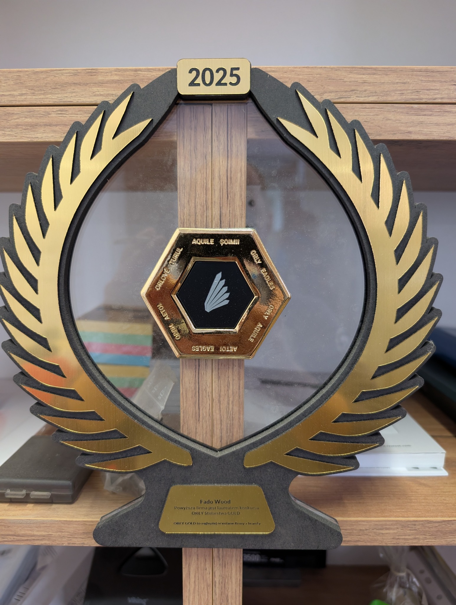 Statuetka Orły Stolarstwa GOLD 2025 dla Fado Wood, z logo w kształcie sześciokąta i złotymi liśćmi laurowymi, stojąca na drewnianej półce.