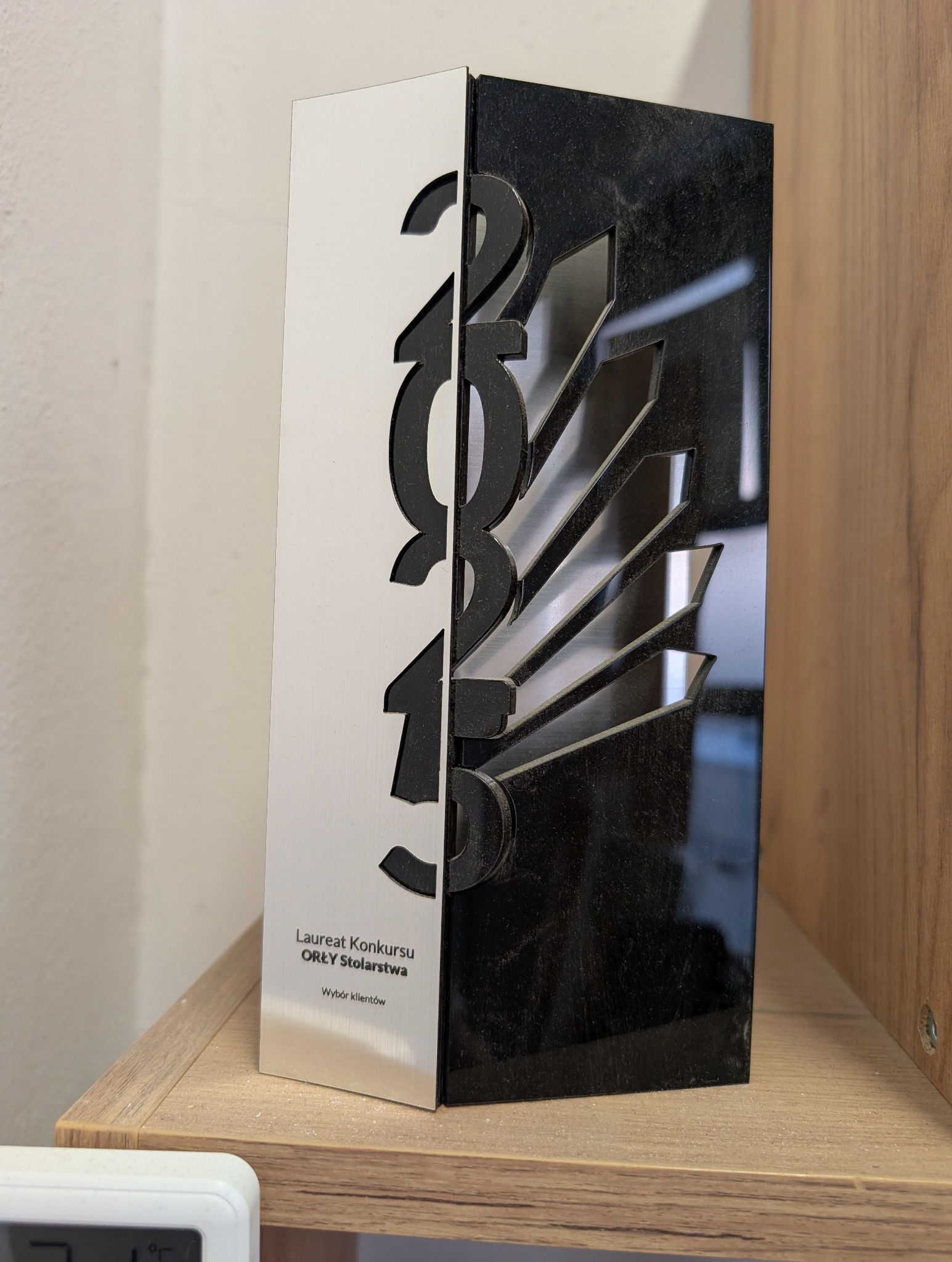 Statuetka 'Orły Stolarstwa' z wyciętym rokiem 2019, stojąca na półce. Nagroda za wybór klientów, w kolorach czerni i bieli, z widocznym kurzem.