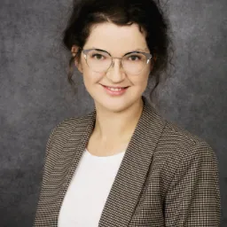 Portret uśmiechniętej kobiety w okularach i brązowej marynarce w kratę na szarym tle.