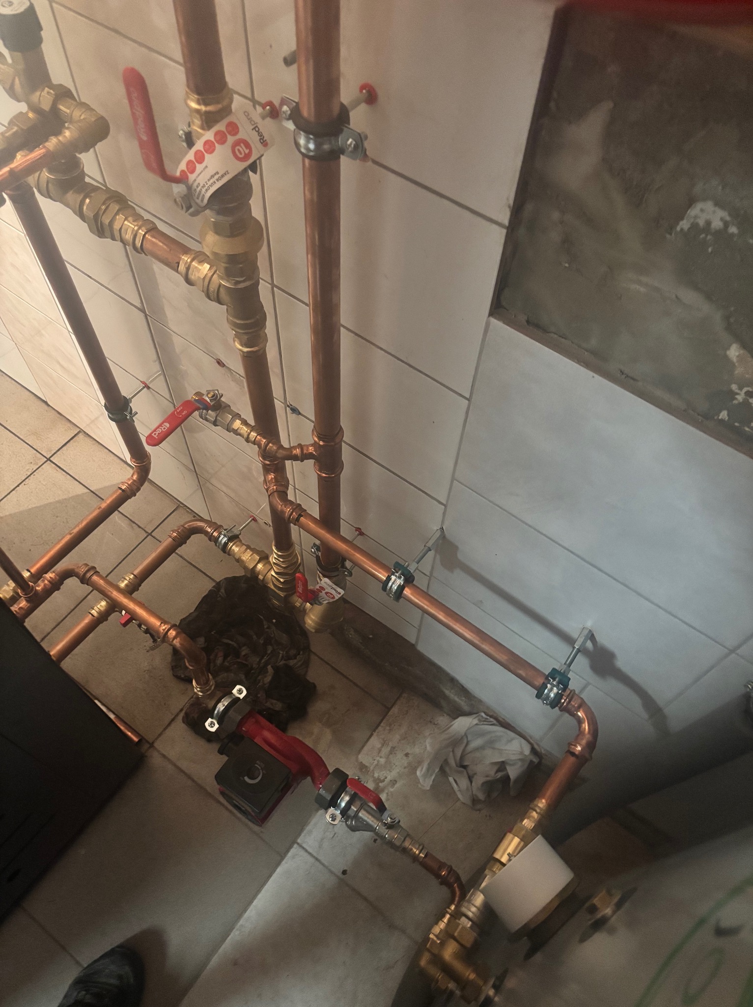 Miedziane rury instalacji grzewczej z zaworami i pompą obiegową, zamontowane przy białej, kafelkowanej ścianie. Widok z góry, ukośny kadr.