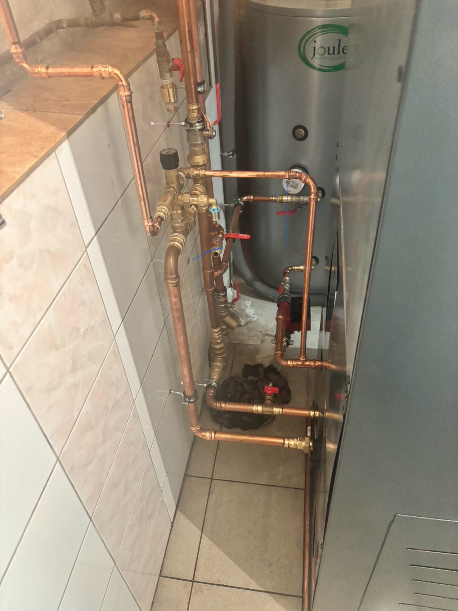 Miedziane rury instalacji CO z zaworami, połączone z srebrnym zasobnikiem Joule, na tle białych i beżowych płytek. Widoczne elementy pompy obiegowej.