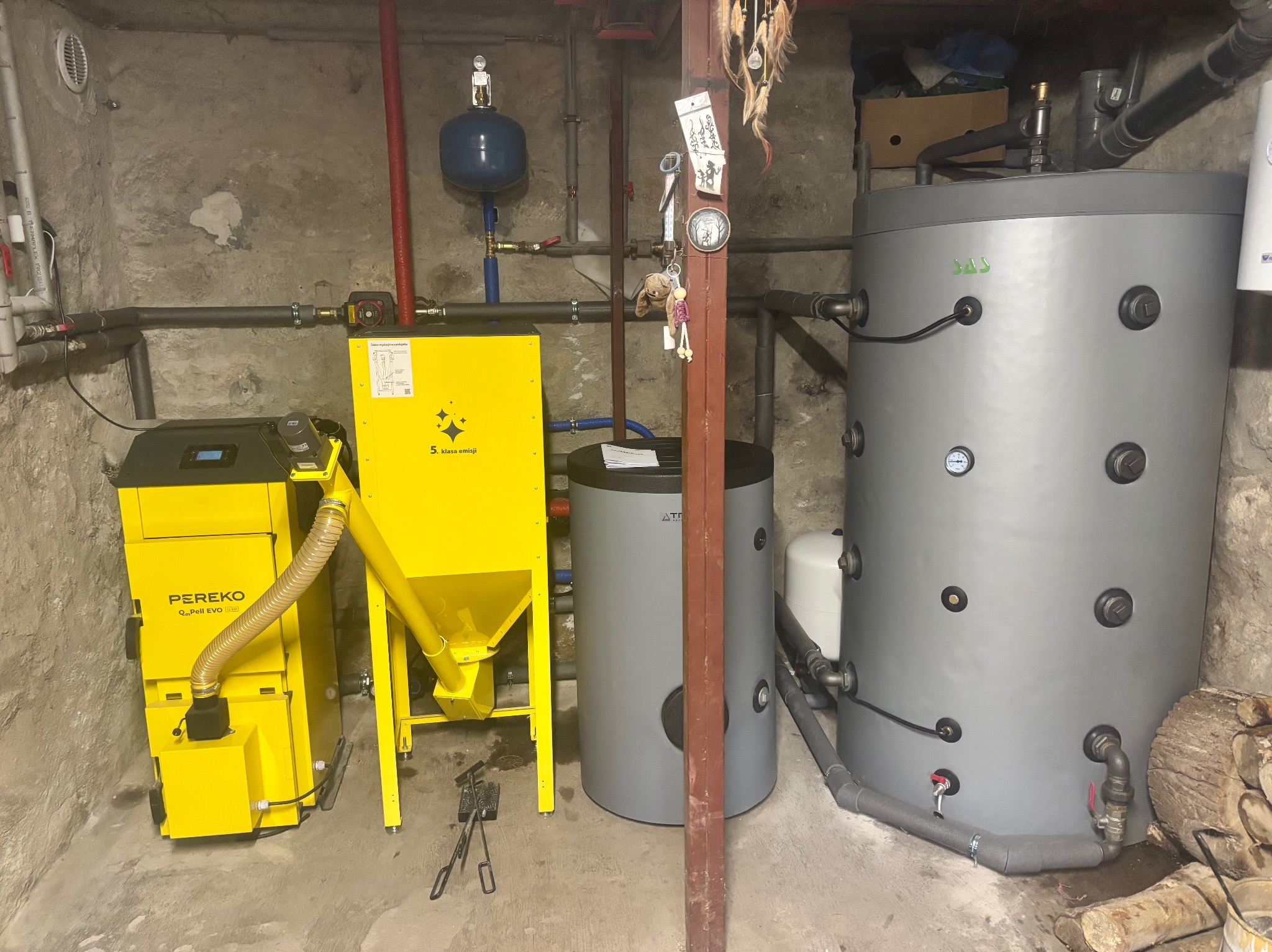 Żółty piec na pellet Pereko Q-Pell EVO z podajnikiem, zbiorniki szare, instalacja CO w piwnicy o surowych ścianach. Widoczne rury, zawory i osprzęt.
