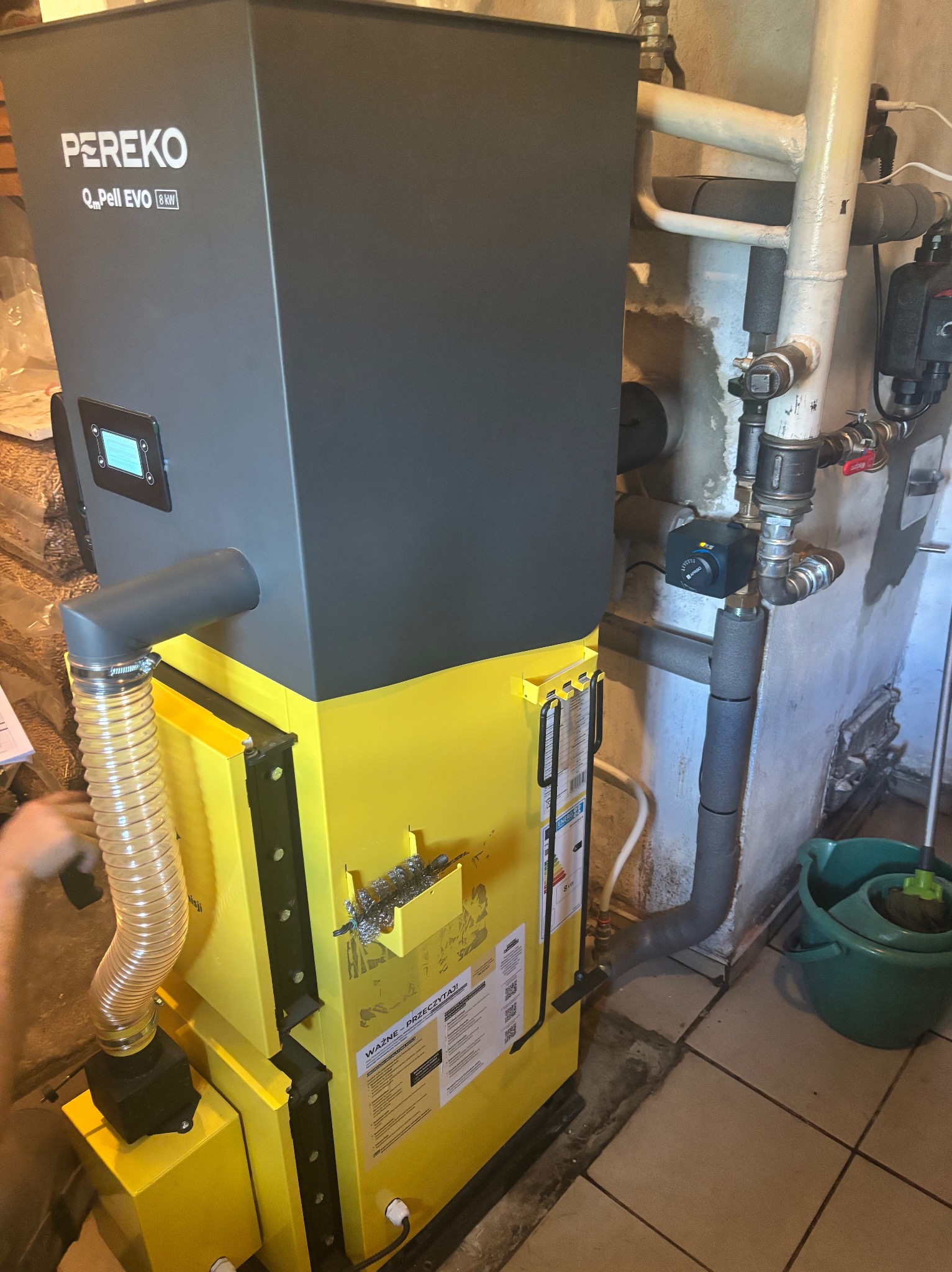 Żółto-szary piec na pellet Pereko Q-Pell EVO 8 kW z podłączonymi rurami w kotłowni, widoczne elementy instalacji grzewczej i fragment podłogi z płytkami.
