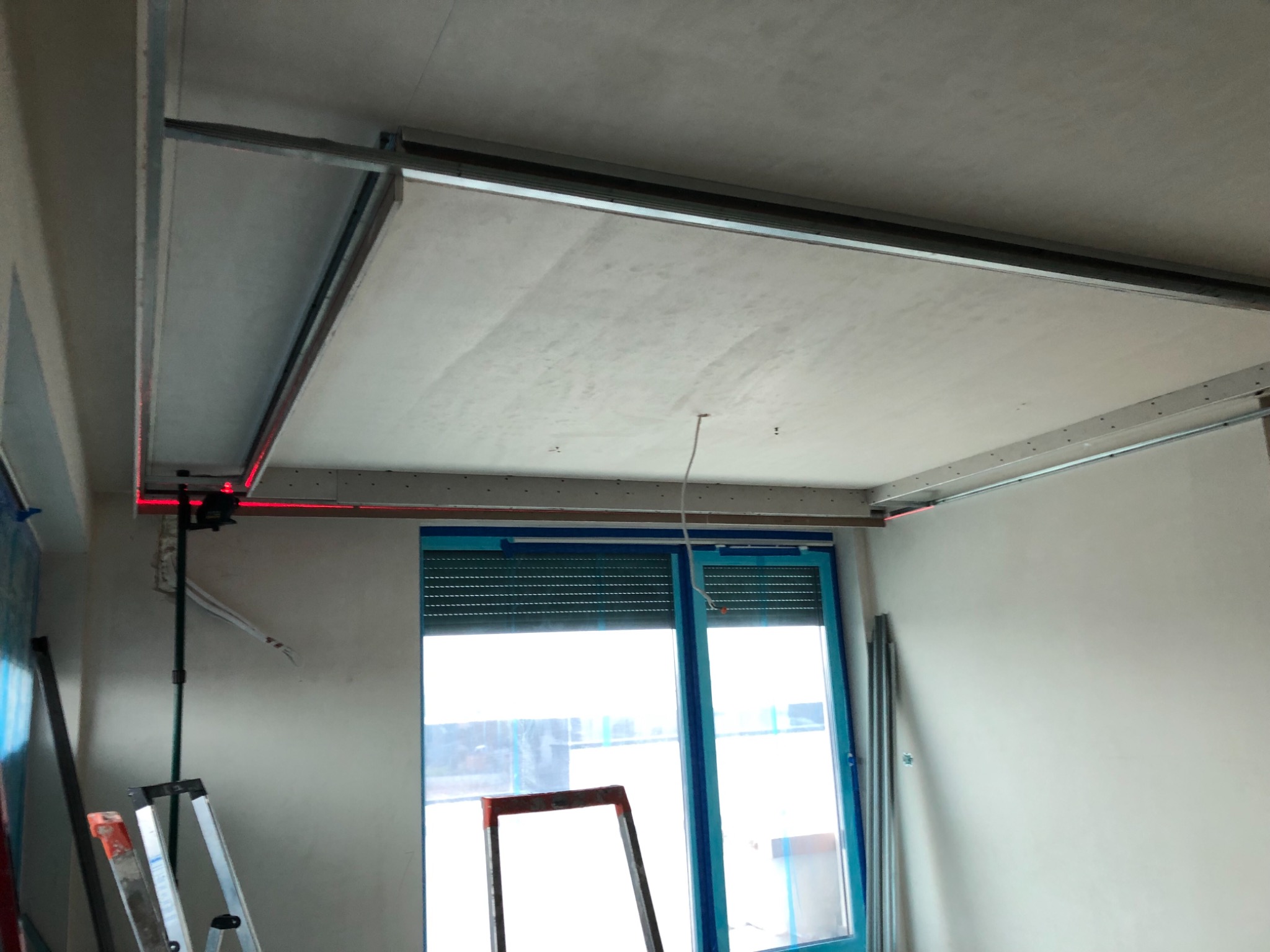 Montaż podwieszanego sufitu z płyt gipsowo-kartonowych, widoczna metalowa konstrukcja i laserowy niwelator, okno zabezpieczone folią.